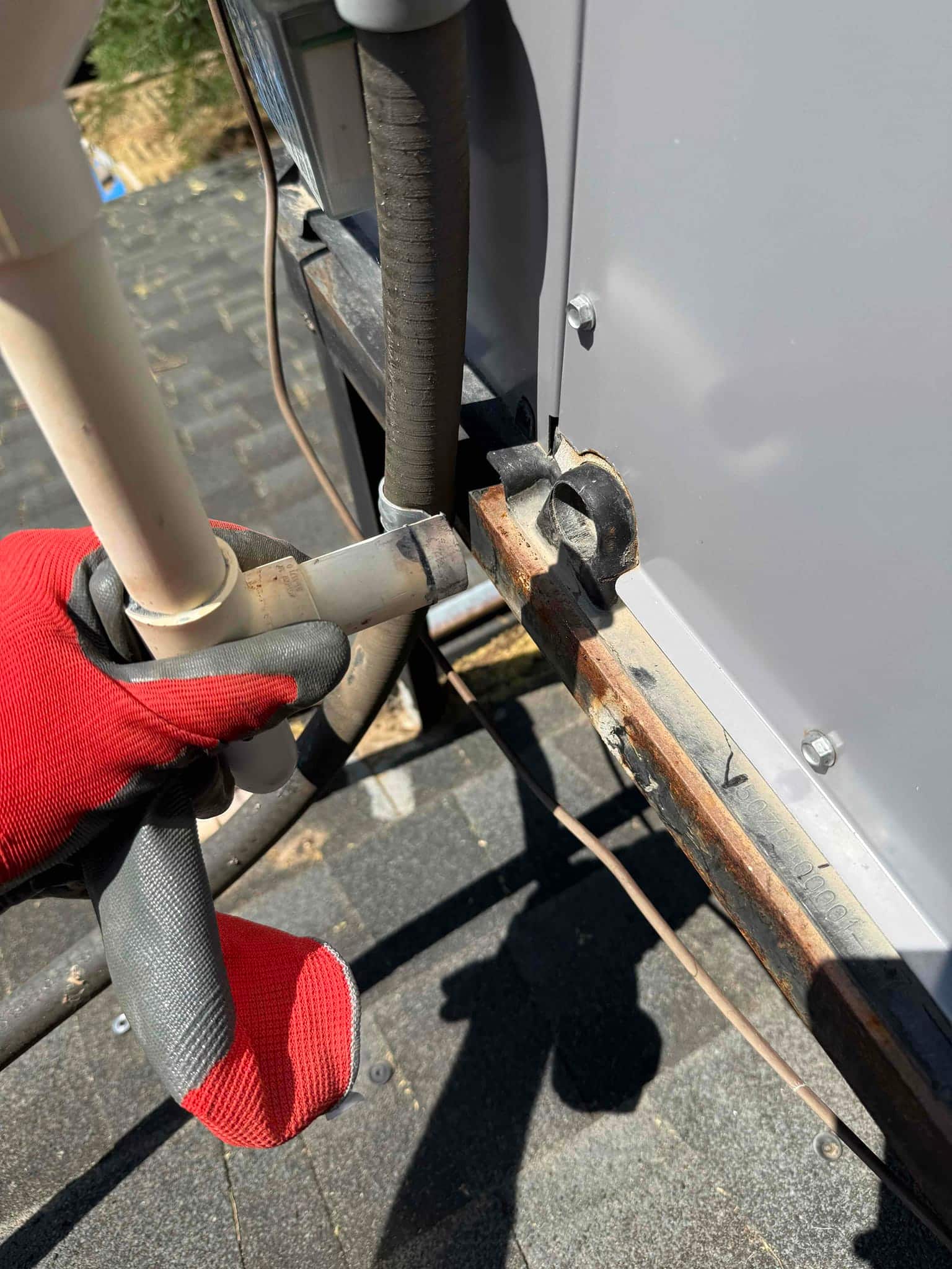 Gloved hand holds PVC condensate drain at the unit’s side near the insulated refrigerant line and rusty mounting rail.
