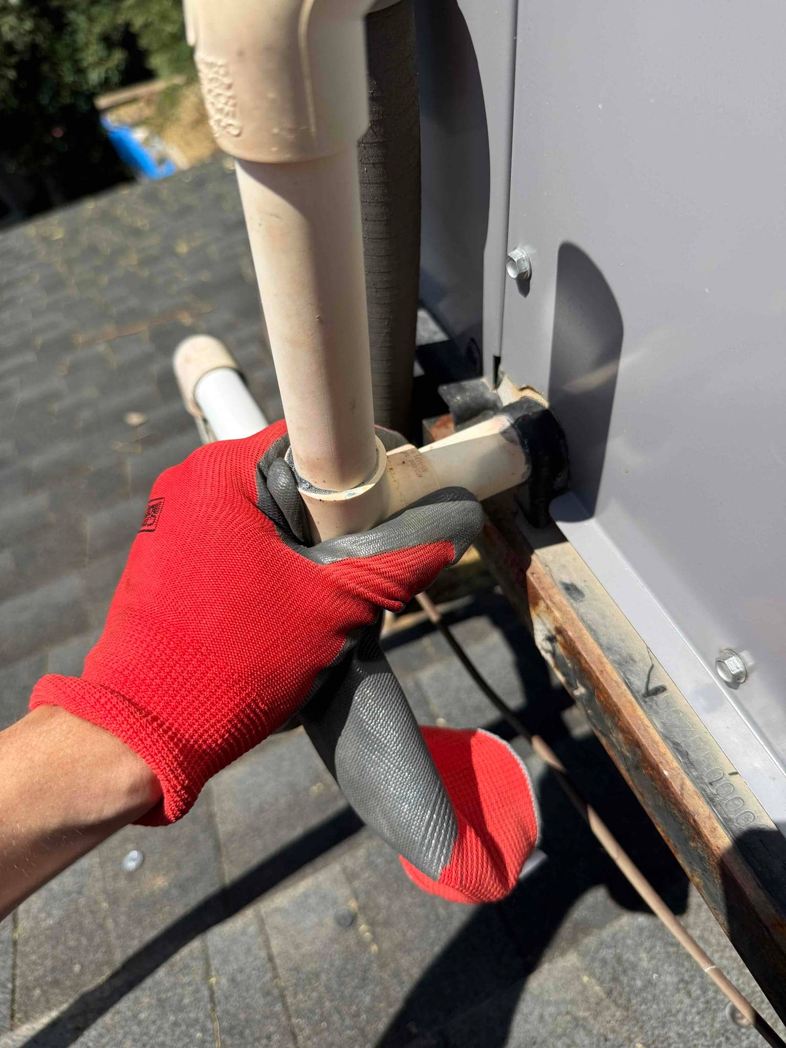 Gloved technician hand holds PVC condensate drain next to insulated refrigerant line at outdoor unit cabinet.
