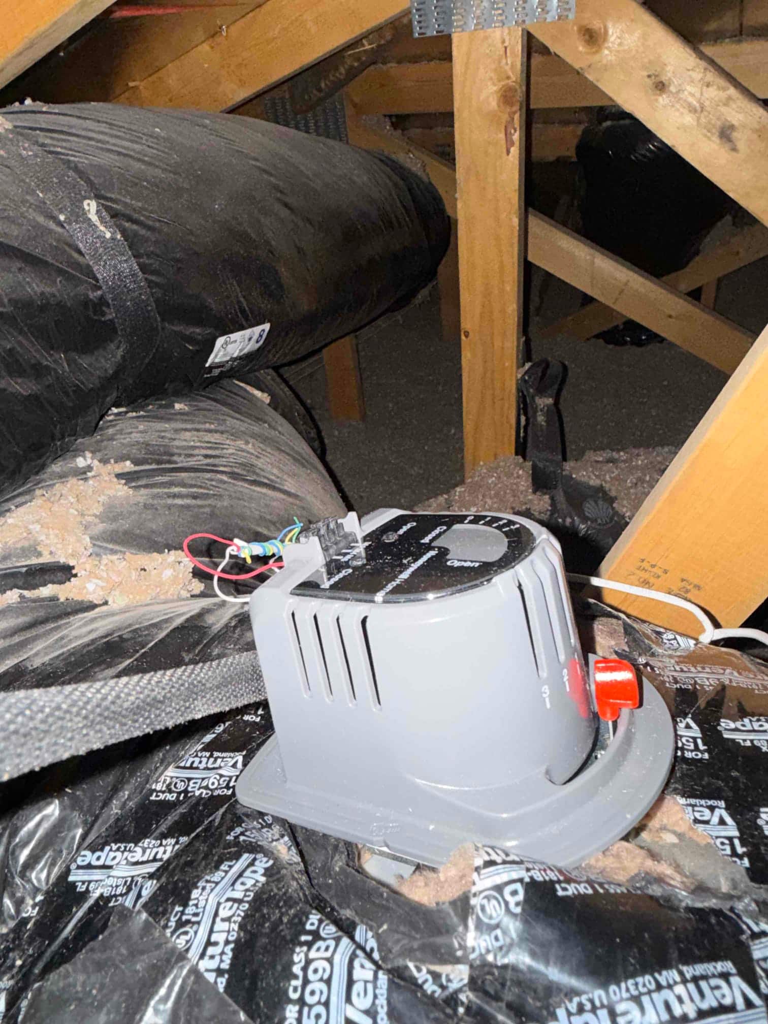 Gray control housing with a dial and red knob rests on attic flexible duct, with low-voltage wires exposed beside it.