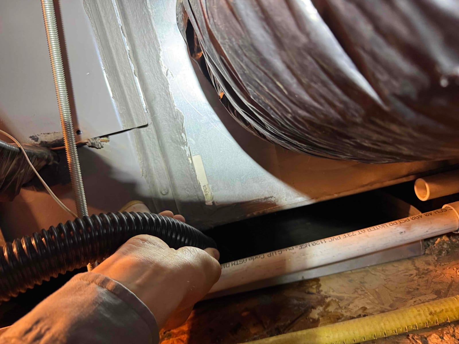 Hand holds a black flexible hose beside the air handler cabinet, with flex duct and PVC condensate drain visible.