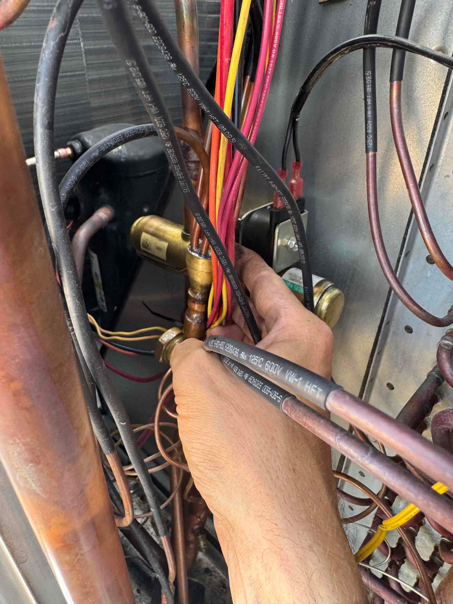 Hand holds bundled control wires near copper refrigerant lines, brass service fitting, and compressor inside an outdoor condenser cabinet.