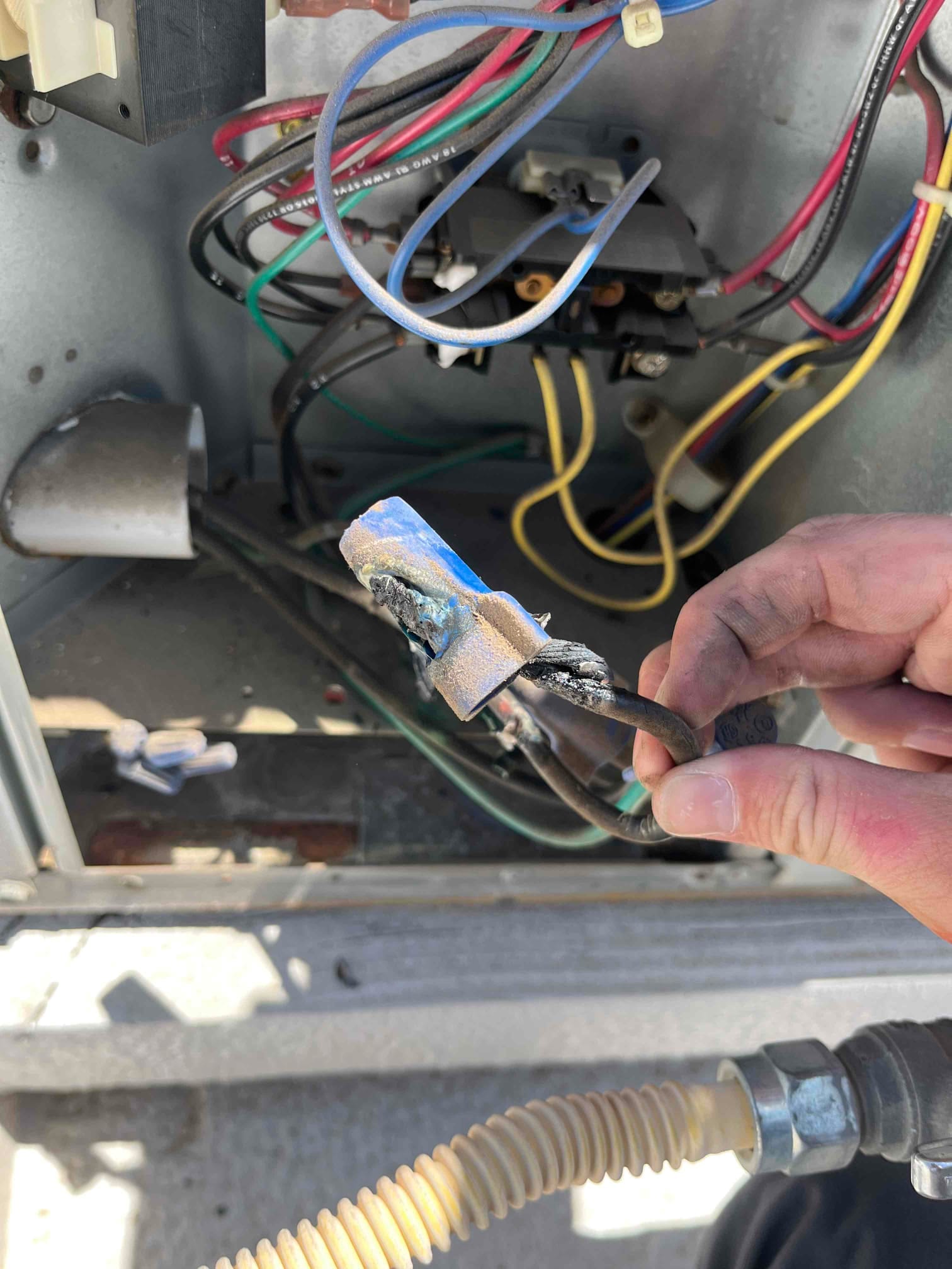 Hand holds a discolored and charred spade connector with attached wires inside an outdoor condenser control compartment.
