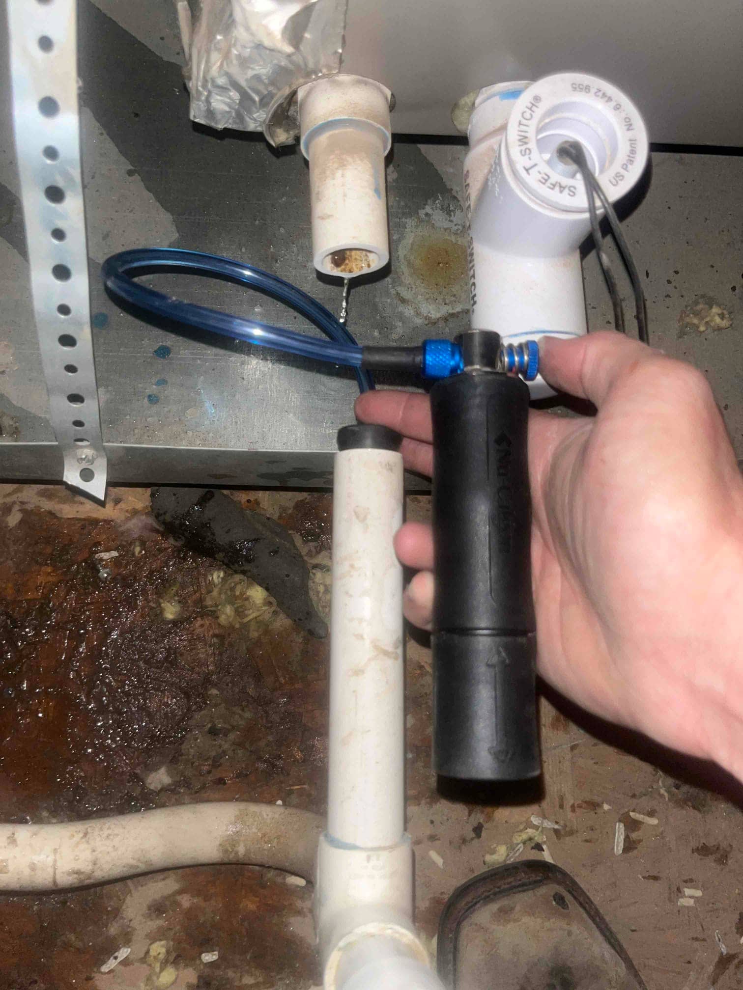 Hand holds a drain line cleaning tool at the PVC condensate piping below the air handler, with a float switch and wet floor.