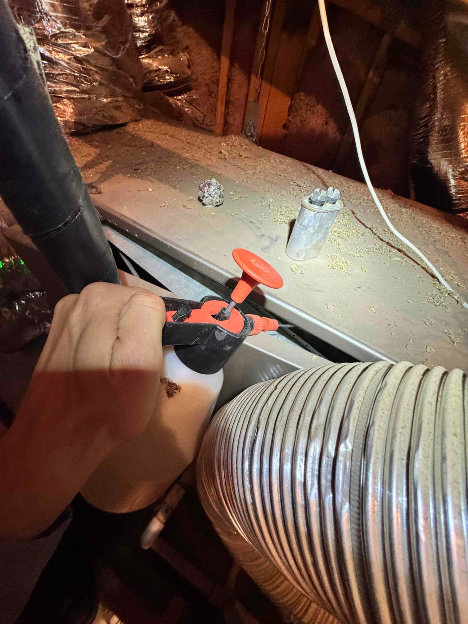 Hand holds a pump sprayer at an attic air handler cabinet beside flexible duct and a two-terminal capacitor.