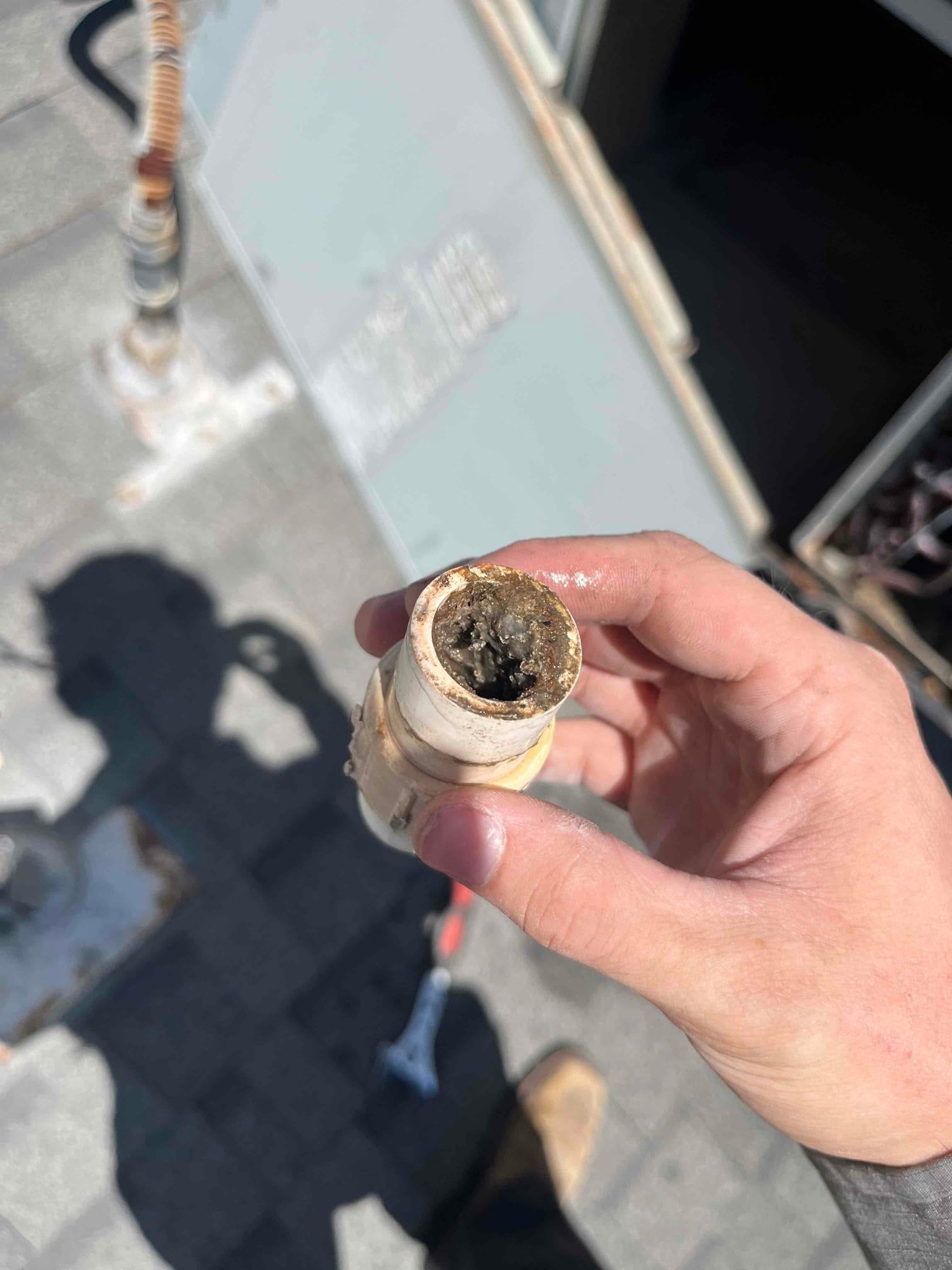 Hand holds a PVC drain fitting packed with dark sludge beside rooftop HVAC unit.