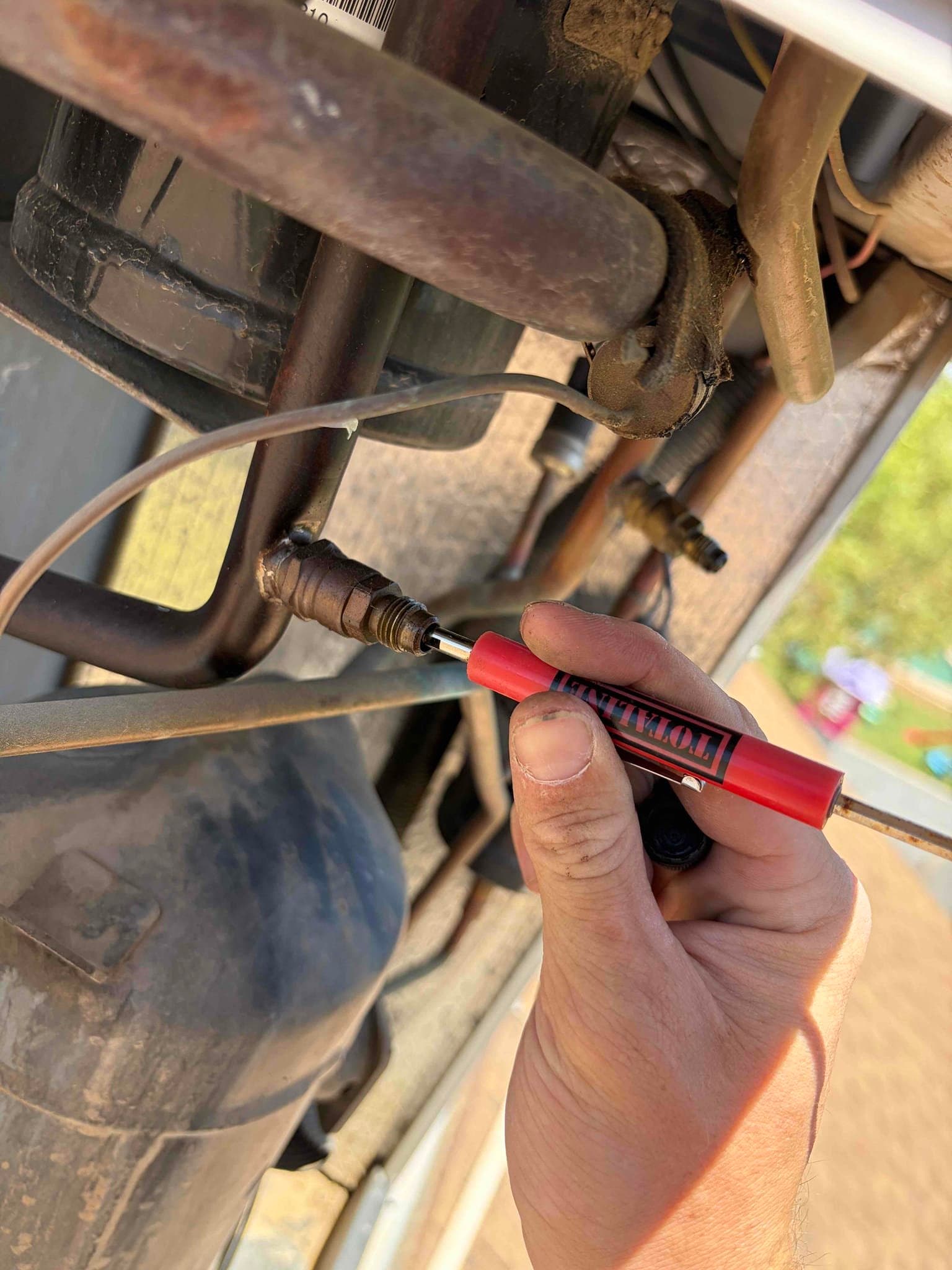 Hand holds a valve core tool on a refrigerant service port beside the condenser compressor tubing.