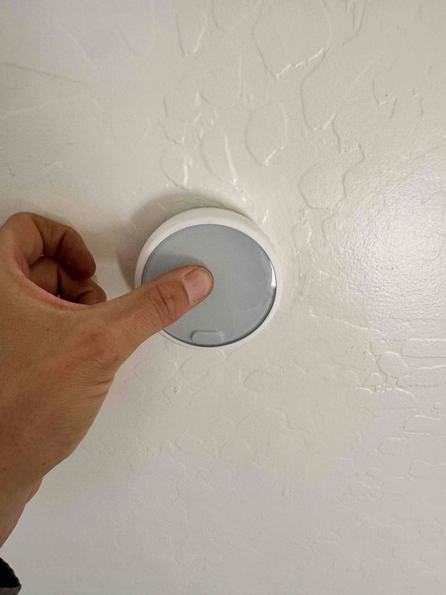 Hand pressing a round wall-mounted thermostat sensor with a smooth gray face and white trim.