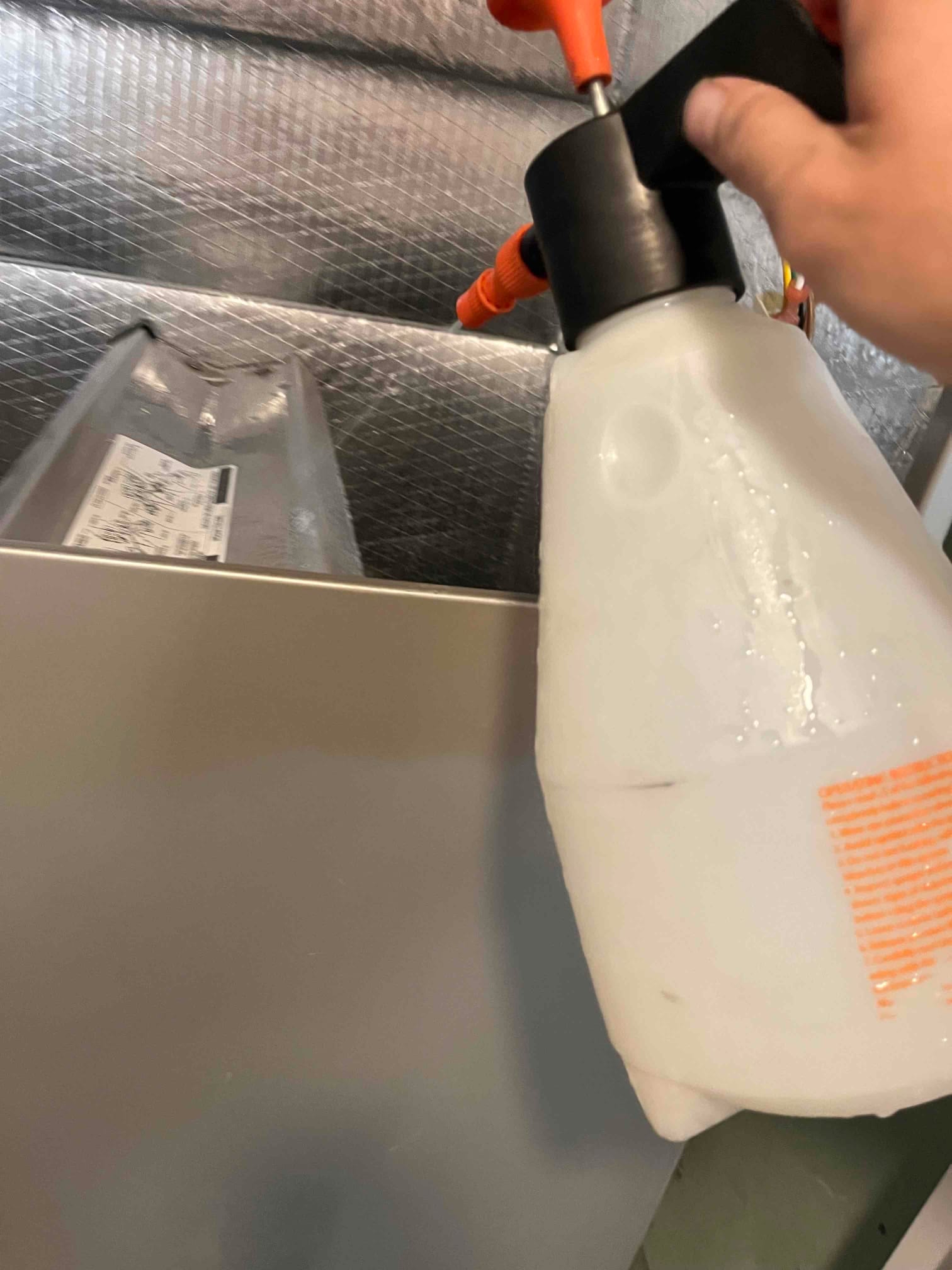 Hand pump sprayer held near an indoor air handler cabinet and insulated ductwork above the unit.