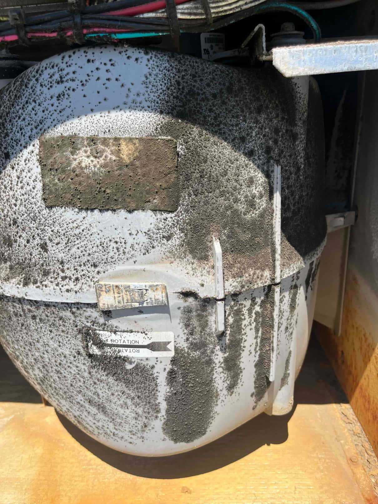 Hermetic compressor shell with heavy surface corrosion and debris, visible rotation arrow label and wiring above.