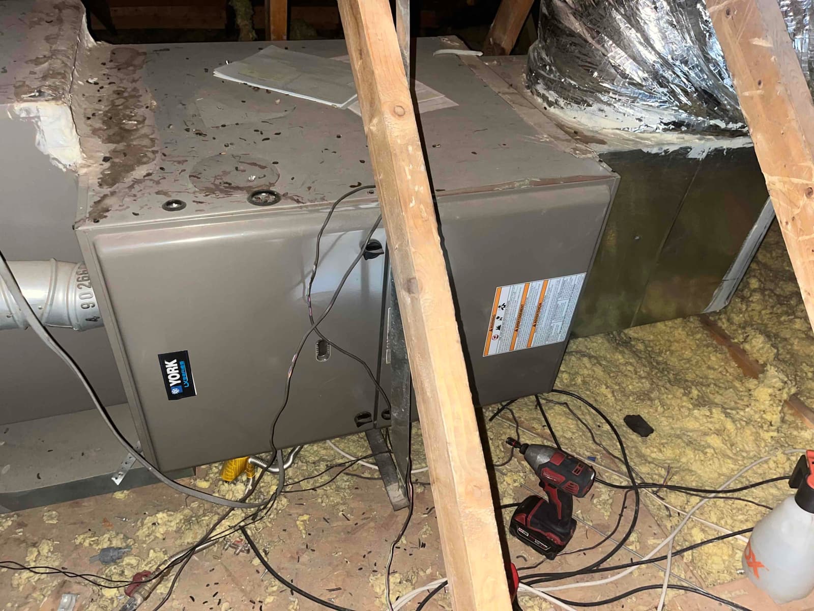 Horizontal furnace in attic with connected ductwork, surface debris on cabinet, loose wiring, and tools on the floor nearby.