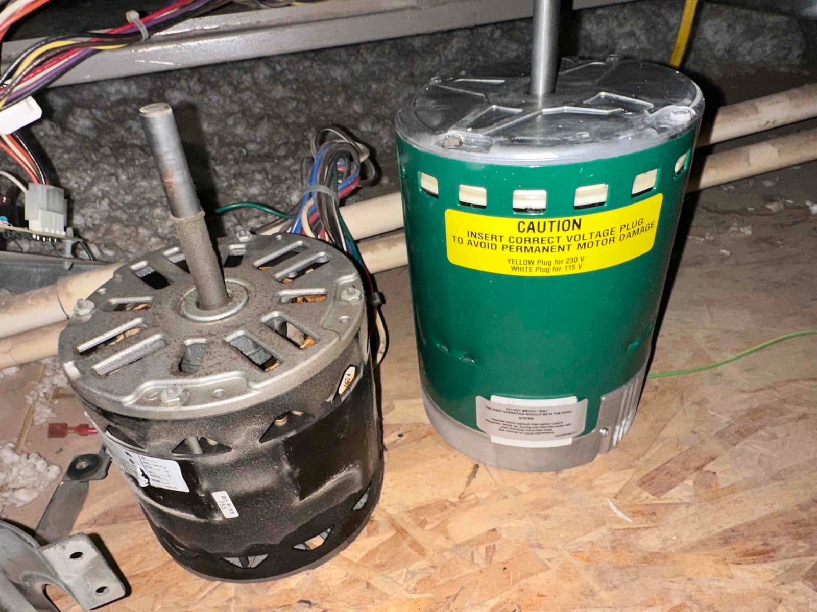 Two HVAC blower motors on the attic floor, dusty black unit beside green motor with visible caution label and wiring.