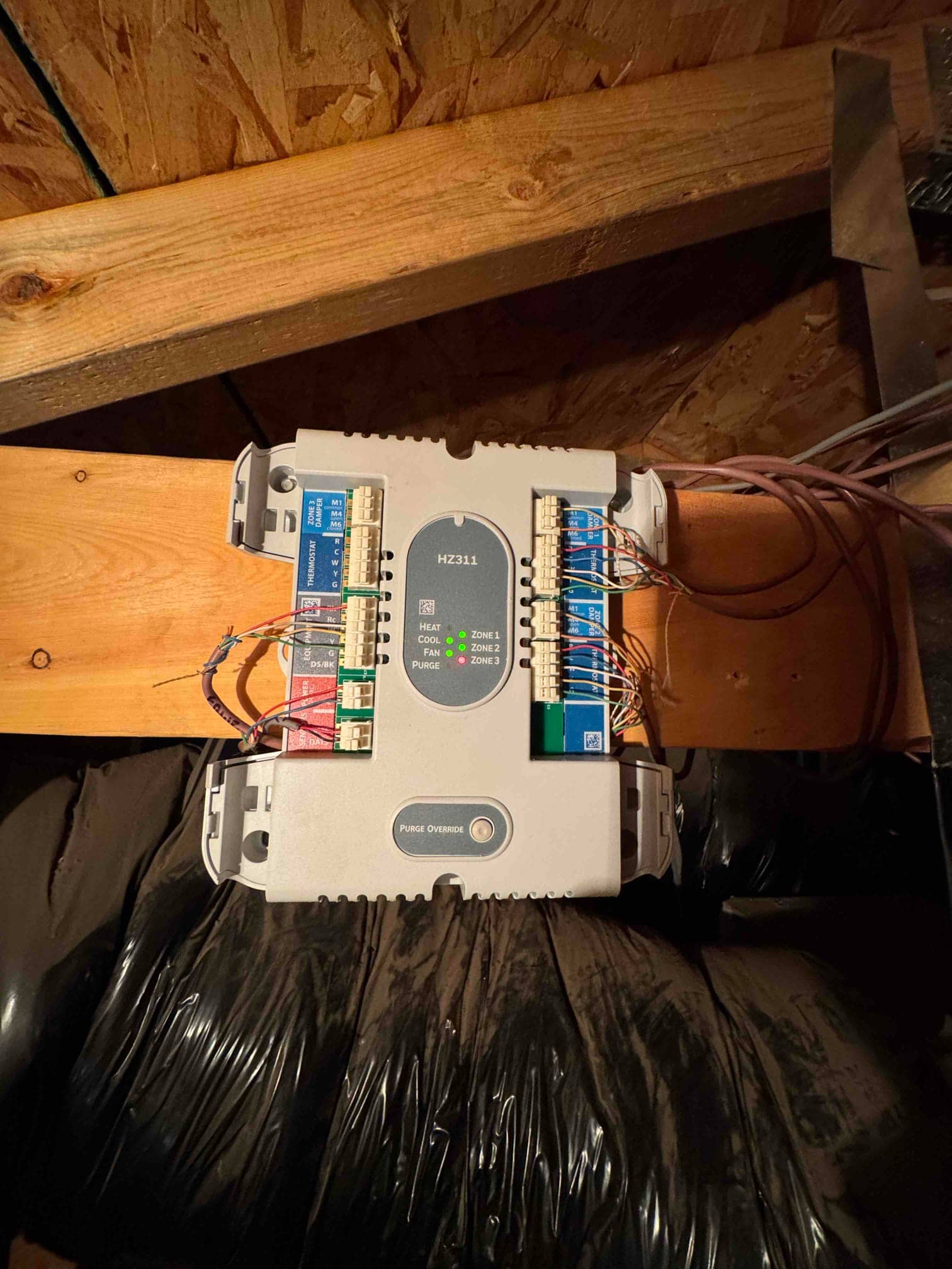 HVAC zoning control board mounted on attic framing, indicator LEDs illuminated, with multiple low-voltage wires connected to zone terminals and purge override button.