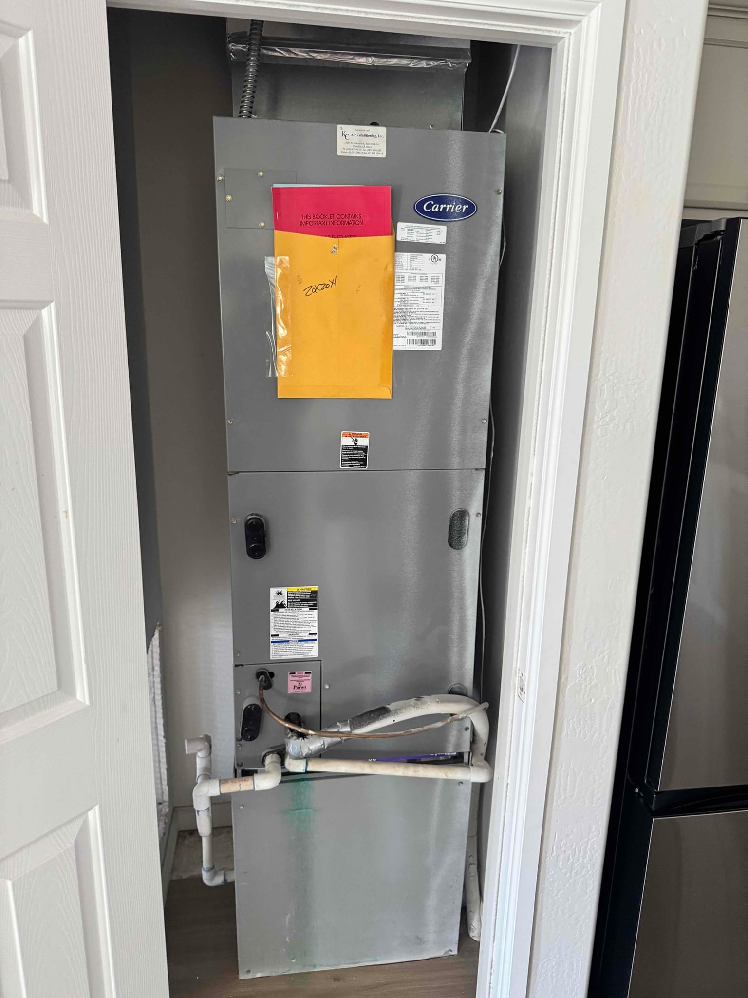 Indoor air handler in closet with PVC condensate drain, insulated refrigerant lines, and service tags attached to cabinet.
