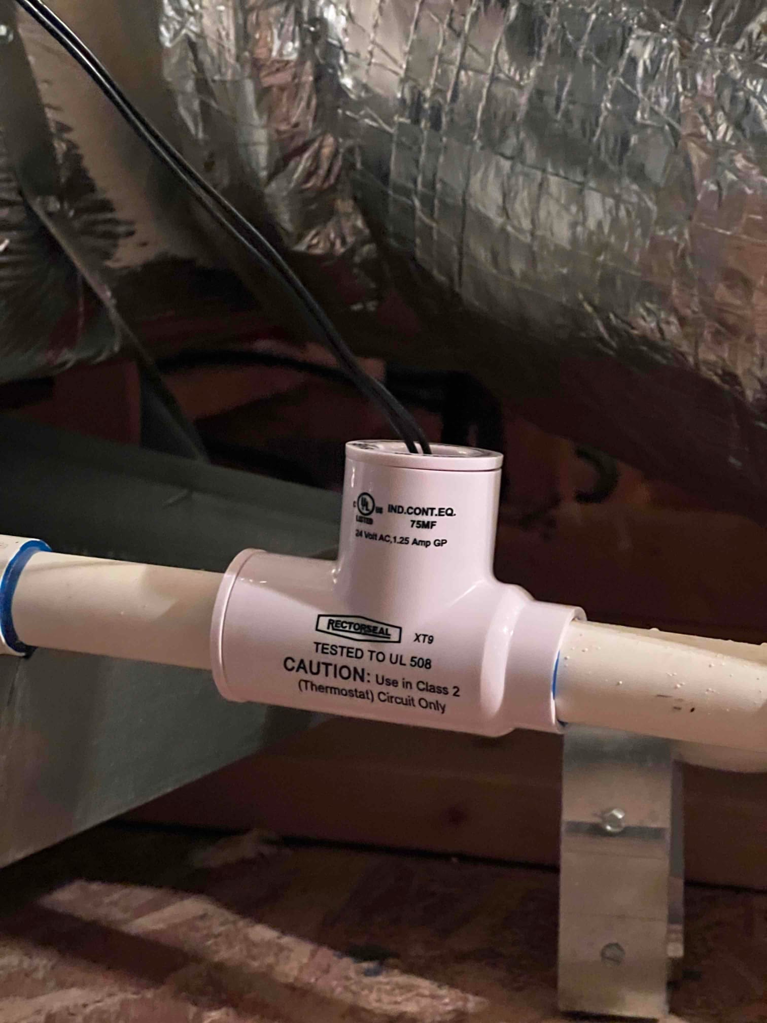 Inline condensate float switch installed on PVC drain with wiring connected and pipe supported by metal strap.