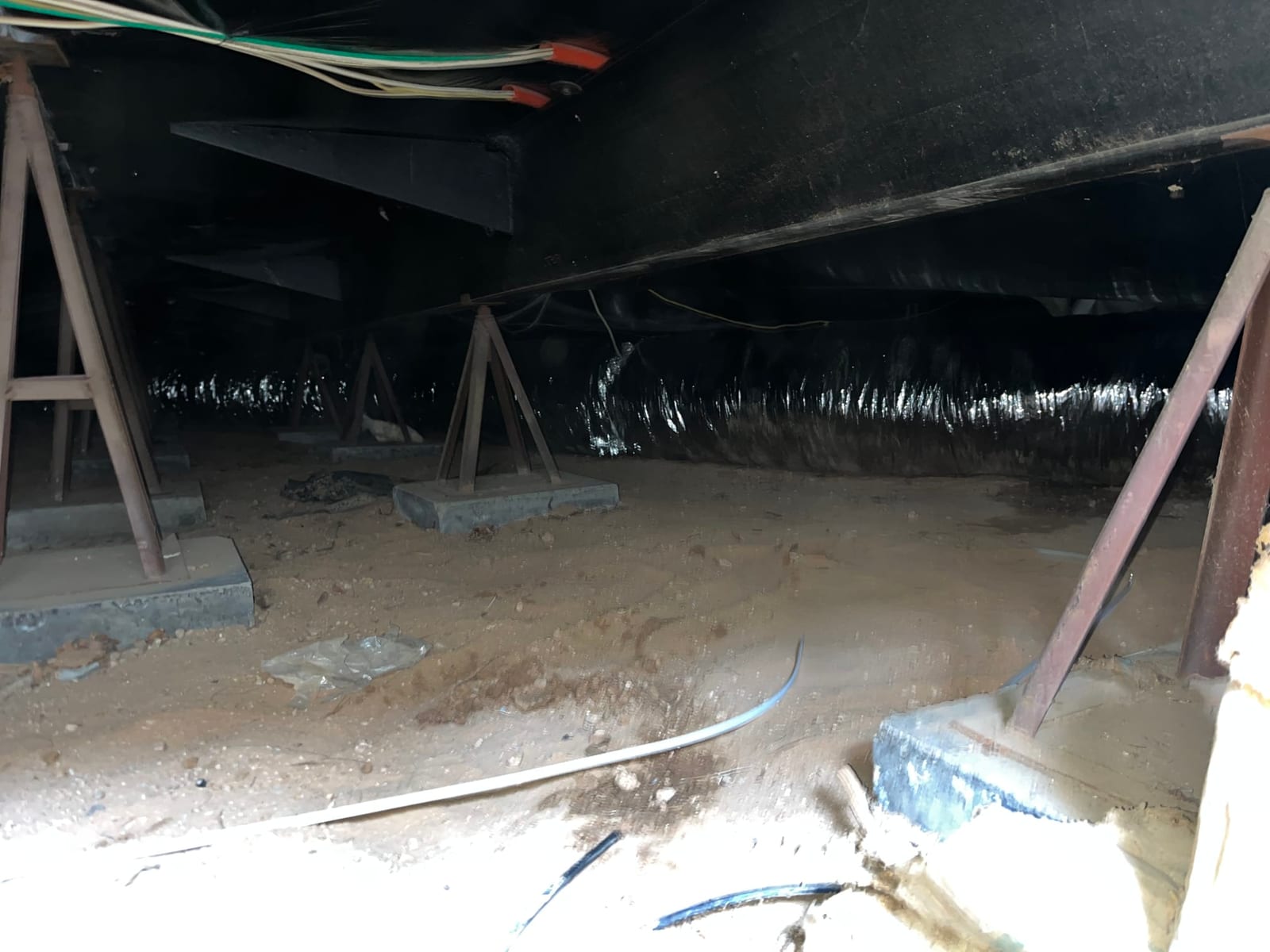 Insulated flexible air duct runs along the crawlspace, with wiring overhead and support piers visible on the sandy floor.
