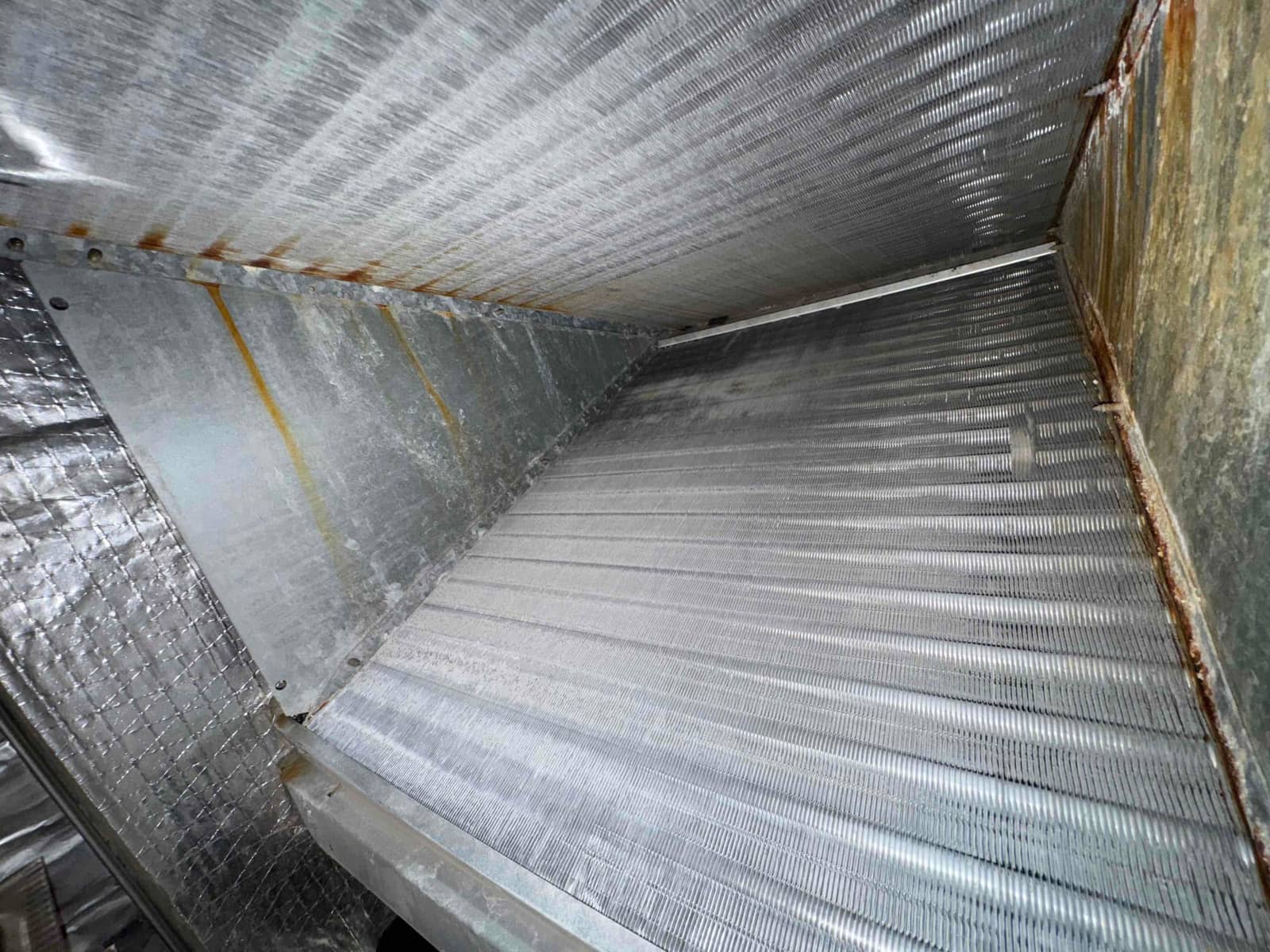 Interior view of an evaporator coil section showing aluminum fins and rust staining along the metal seams.