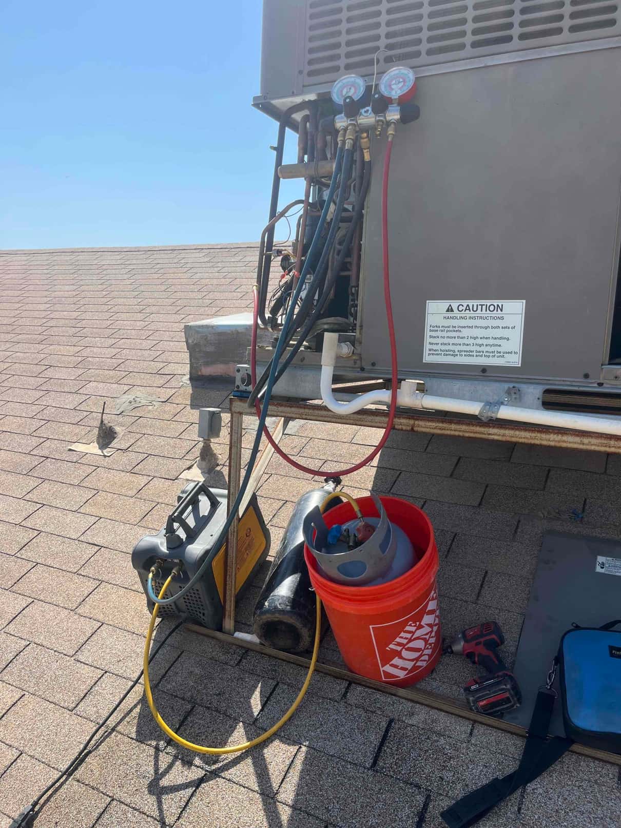 Manifold gauges and hoses connect to a rooftop HVAC unit, with a refrigerant cylinder, bucket, and cordless tool nearby.