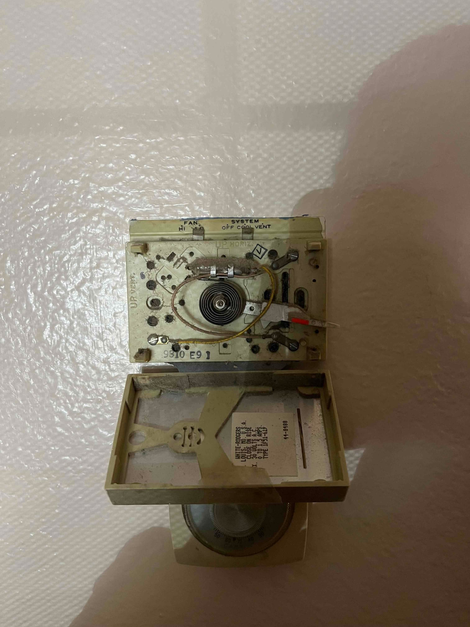 Mechanical wall thermostat with cover open, showing bimetal coil, wiring, and fan/system switch labels.
