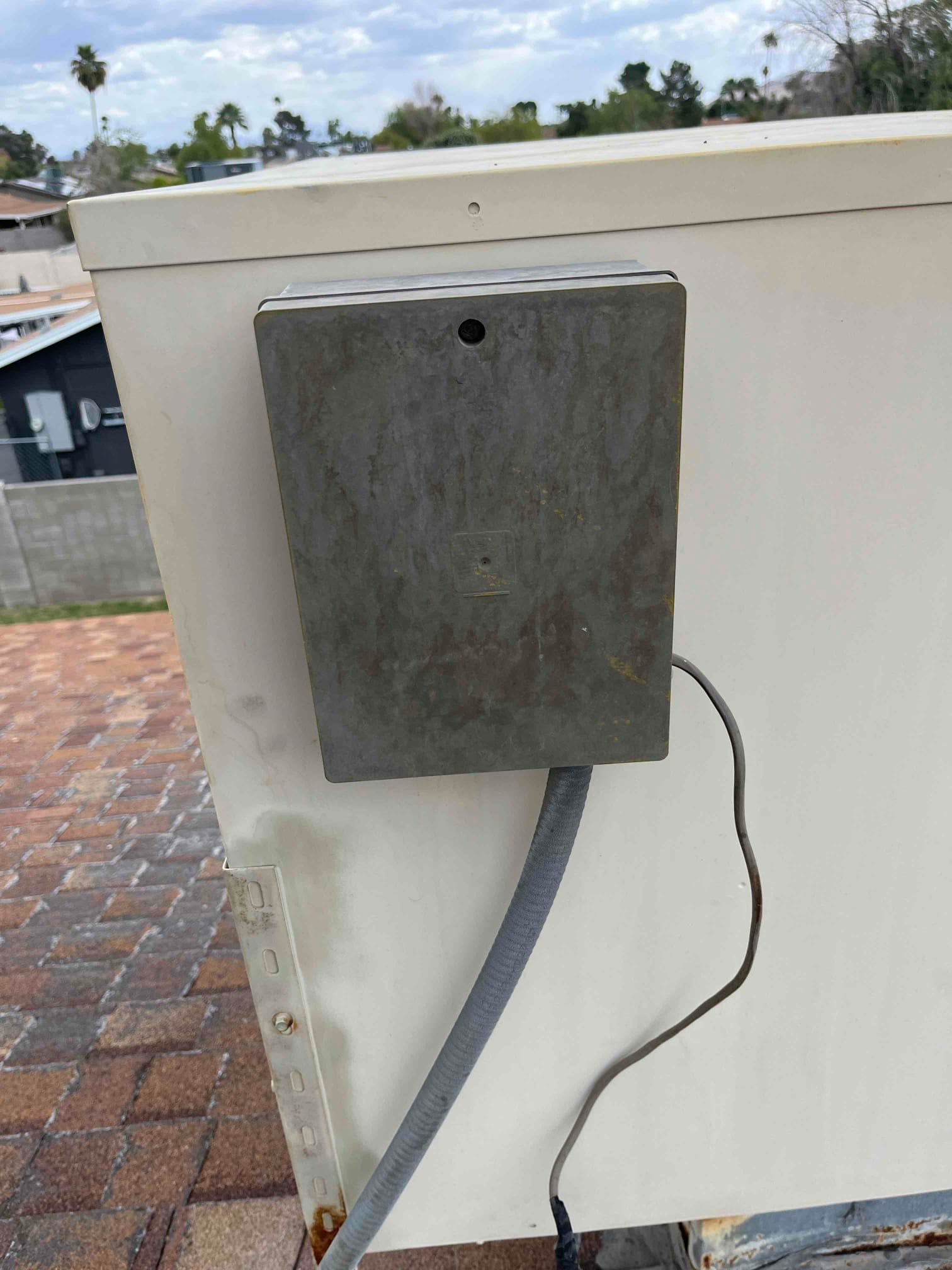 Metal disconnect box mounted on rooftop HVAC unit, with flexible electrical conduit and a separate thin cable.