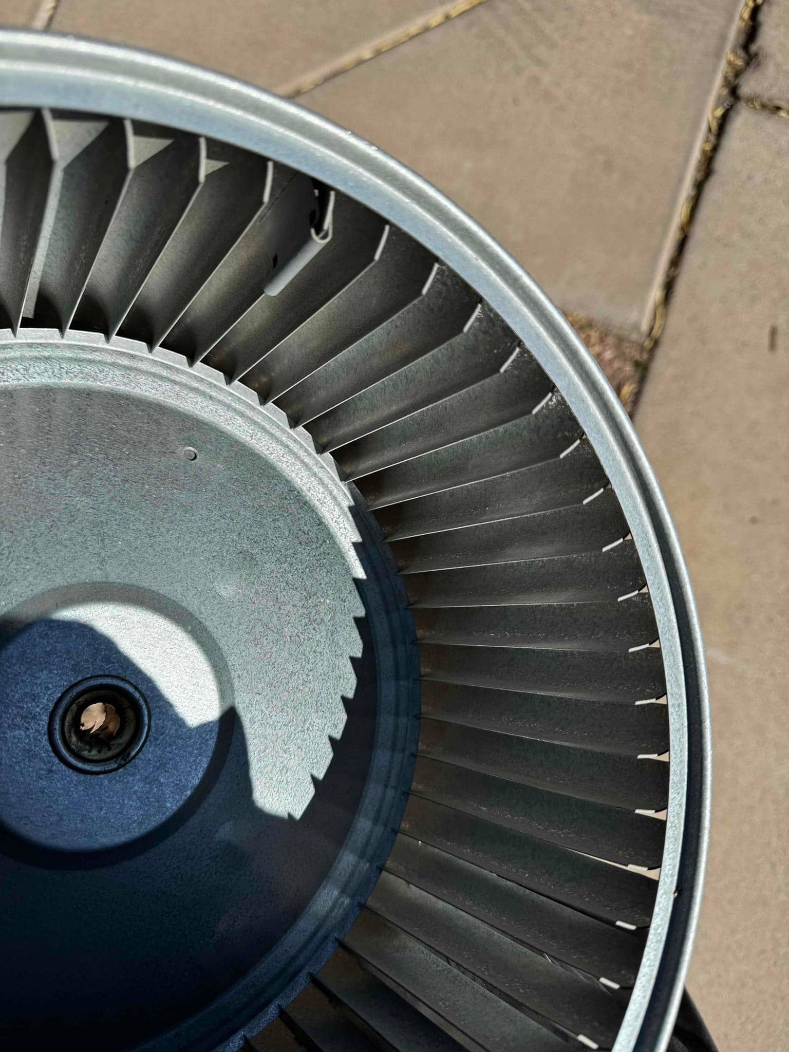Metal HVAC blower wheel with clean radial fins and central hub, removed from housing and resting on pavement.