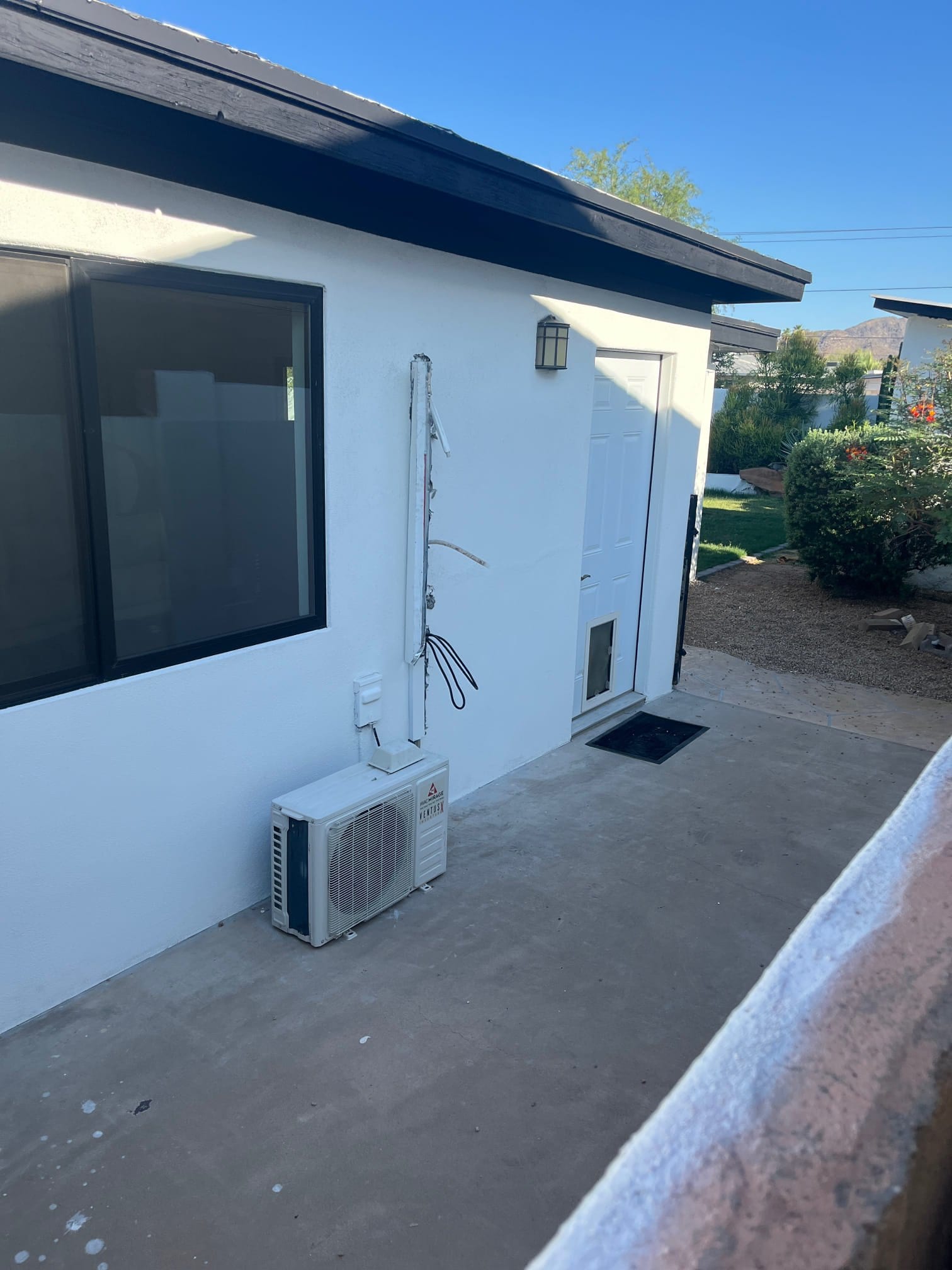 Mini-split outdoor condenser on patio beside wall, with line set cover open and exposed refrigerant lines and wiring.