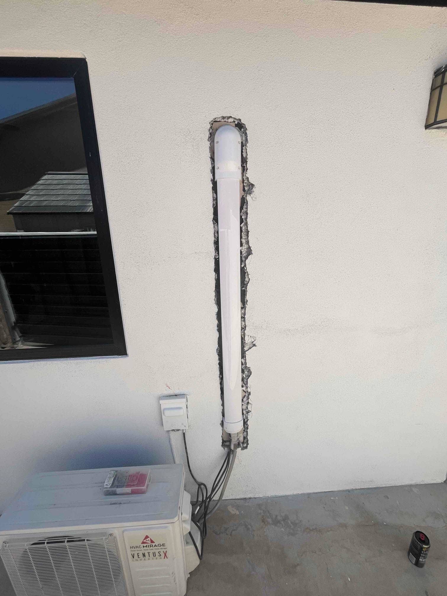 Mini-split outdoor condenser sits below a vertical line-set cover recessed into the wall, with refrigerant lines and an electrical disconnect visible.