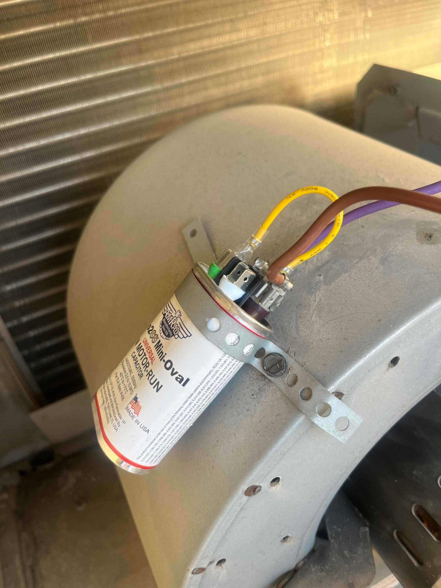 Motor run capacitor strapped to the blower housing, with yellow, brown, and purple wires attached to its terminals.