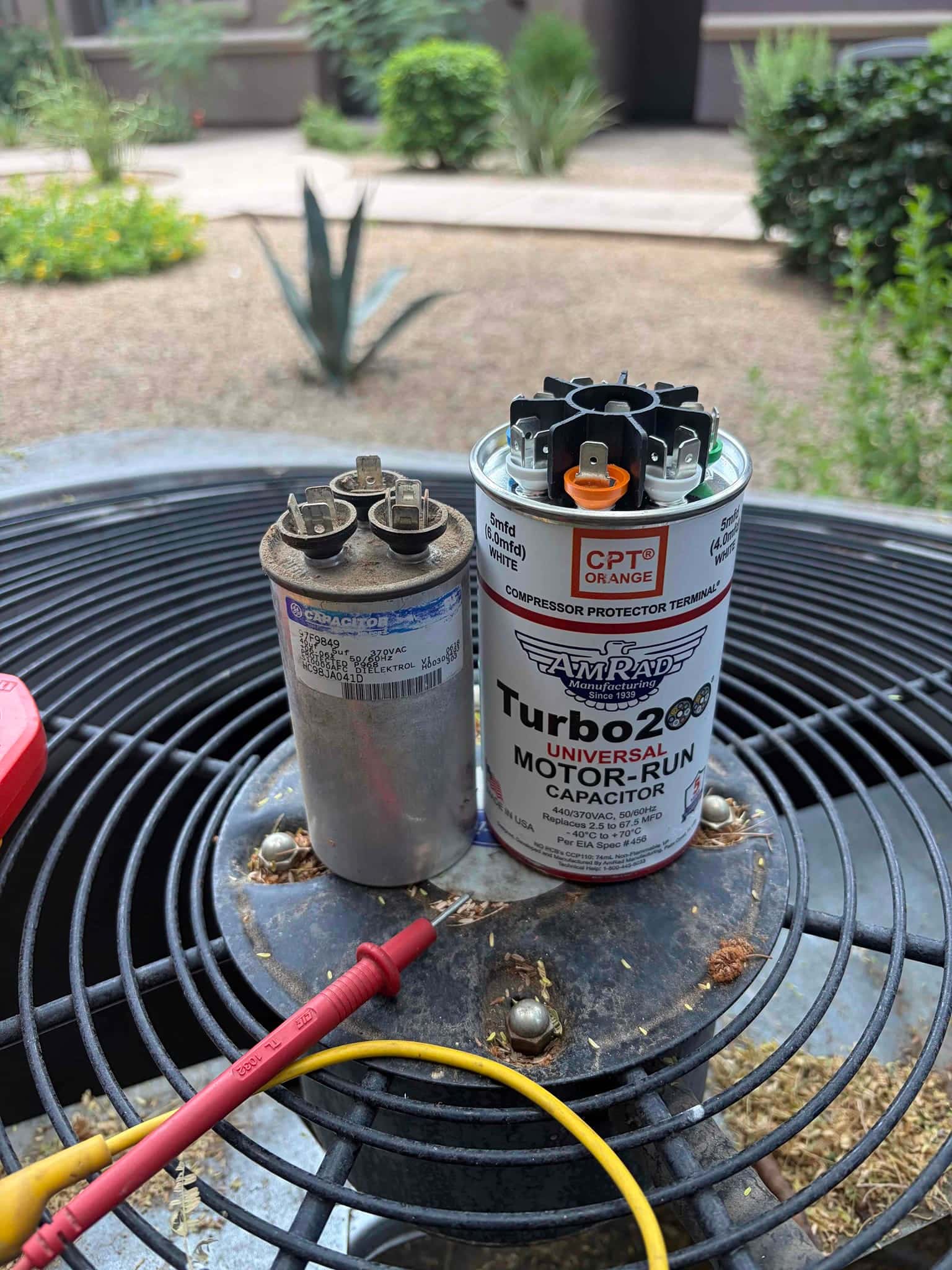 Two motor-run capacitors sit on the outdoor condenser top, alongside red and yellow test leads.