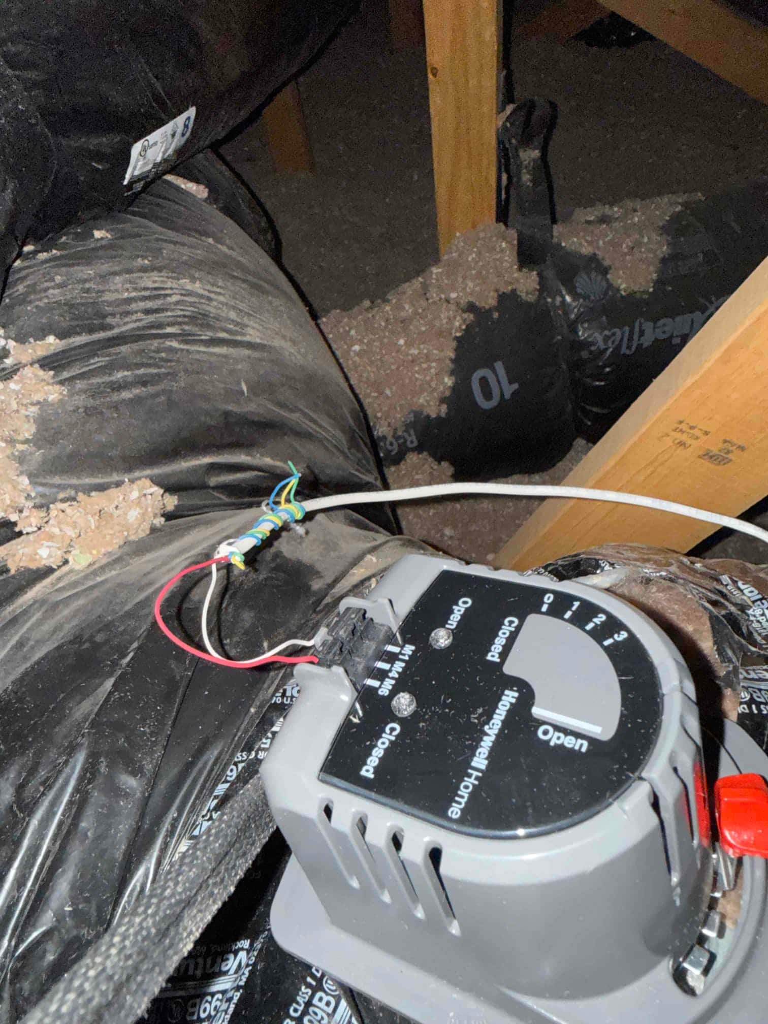 Motorized duct damper actuator mounted on flexible duct in attic, with connected control wires and insulation debris nearby.