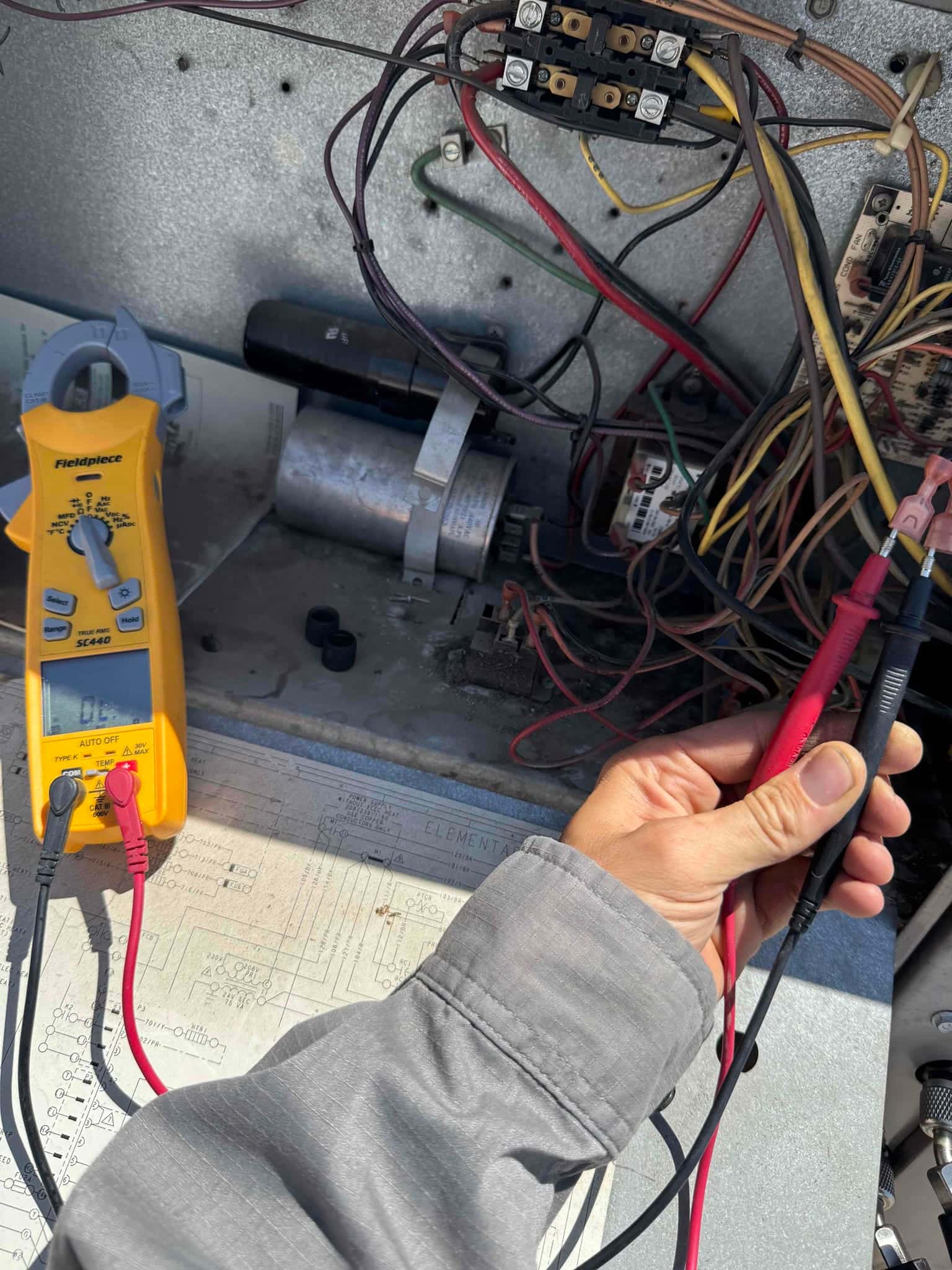 Open condenser control compartment showing wiring, contactor, cylindrical capacitors, and a clamp meter with test leads during electrical checks.
