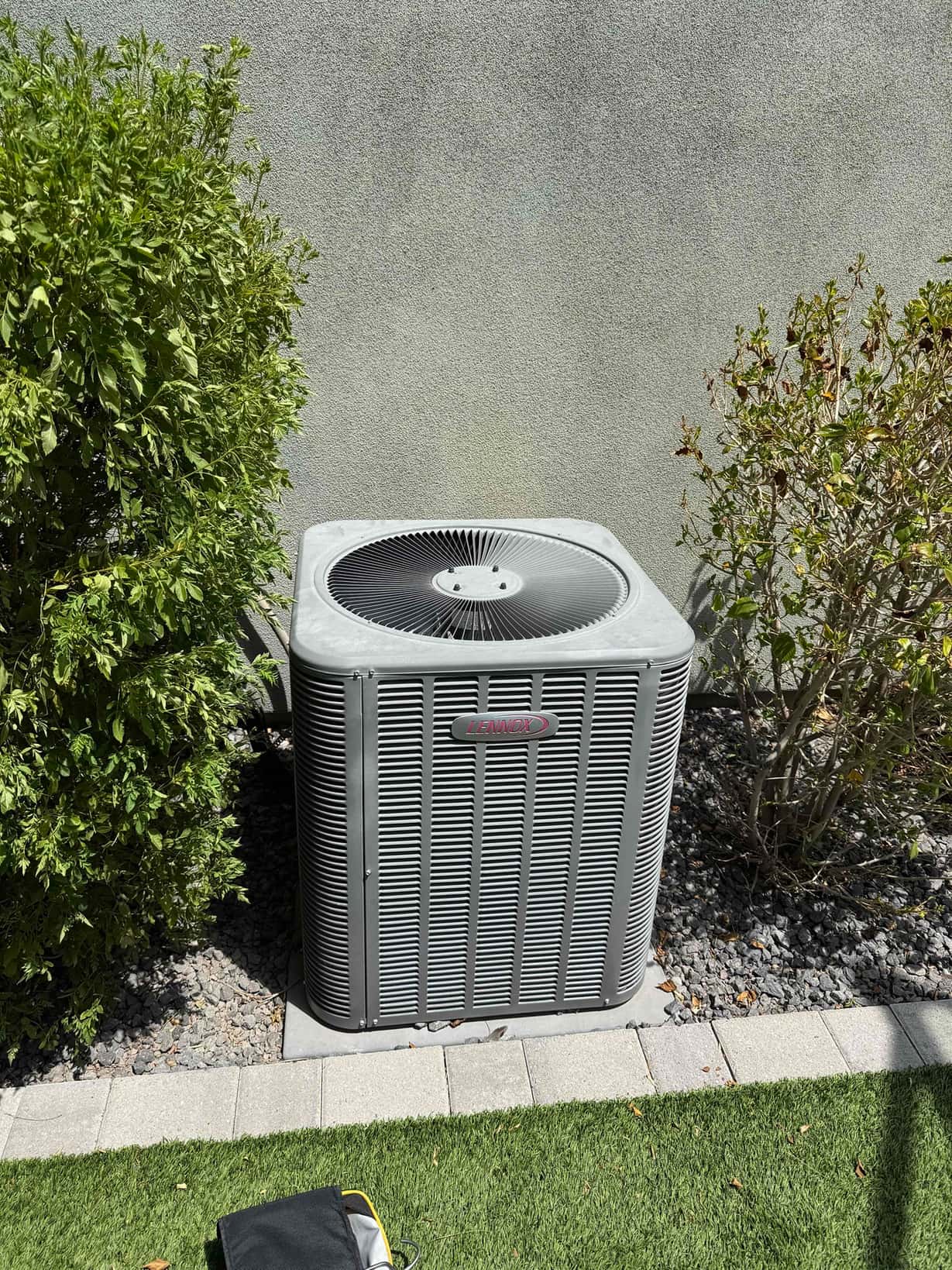 Outdoor air conditioner condenser sits on a pad beside a stucco wall, with shrubs nearby.