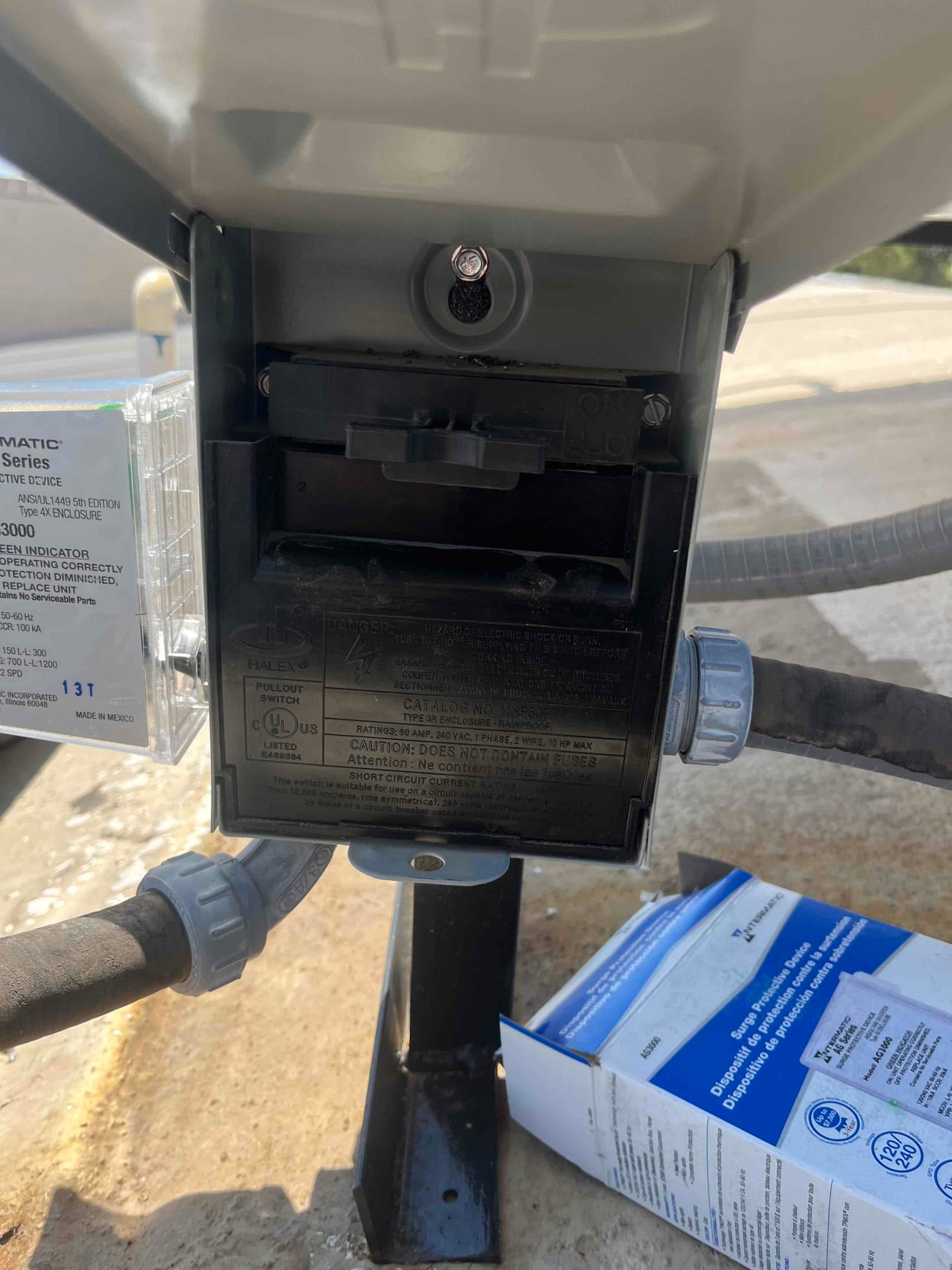 Outdoor air conditioner disconnect with pullout switch open, showing line conduits and mounting bracket, with a surge protective device box nearby.