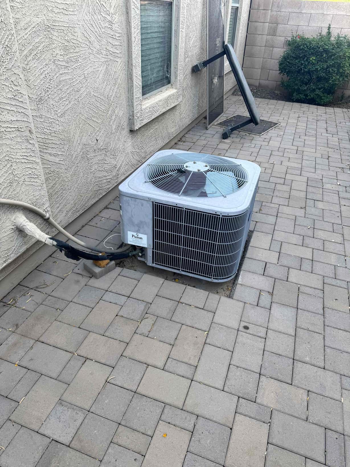 Outdoor air-conditioning condenser beside the house with insulated refrigerant lines and electrical whip on paver patio.