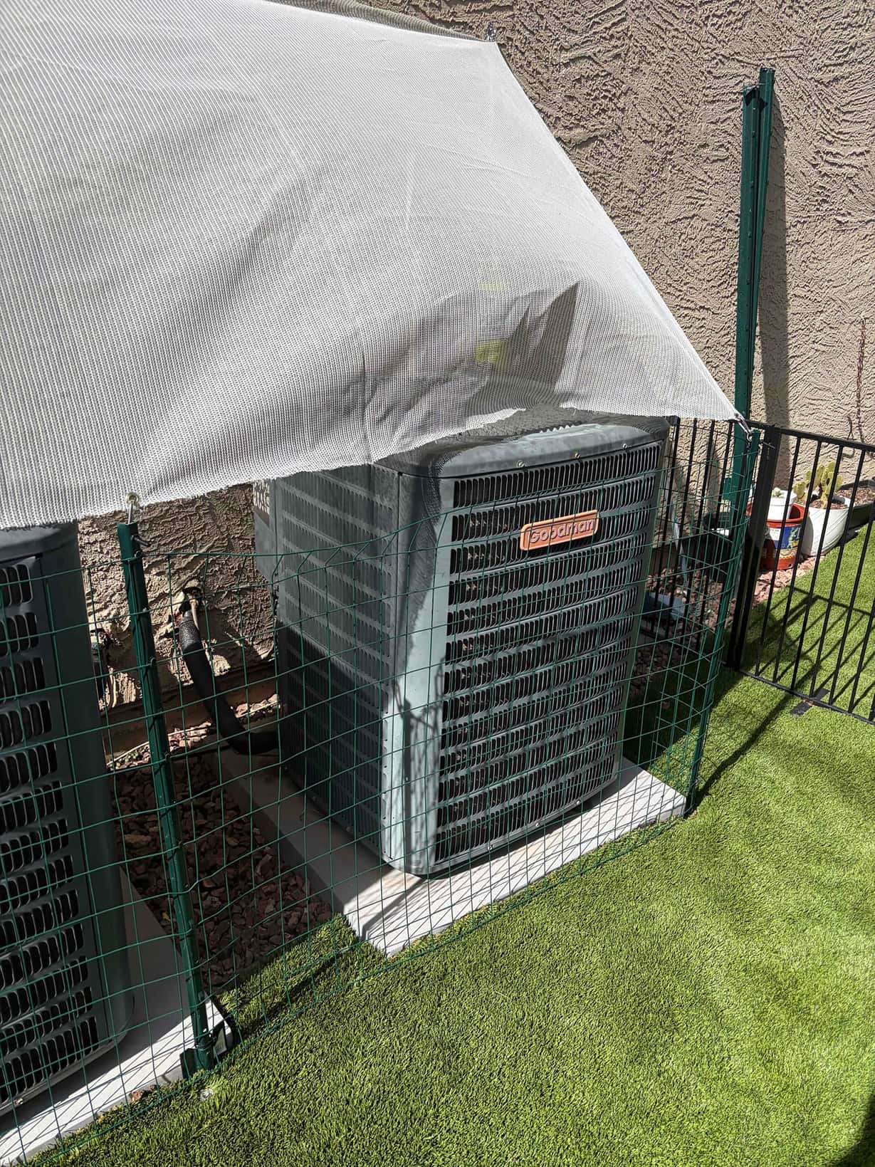 Two outdoor air-conditioning condenser units sit on pads under a mesh sun shade within a protective fence.