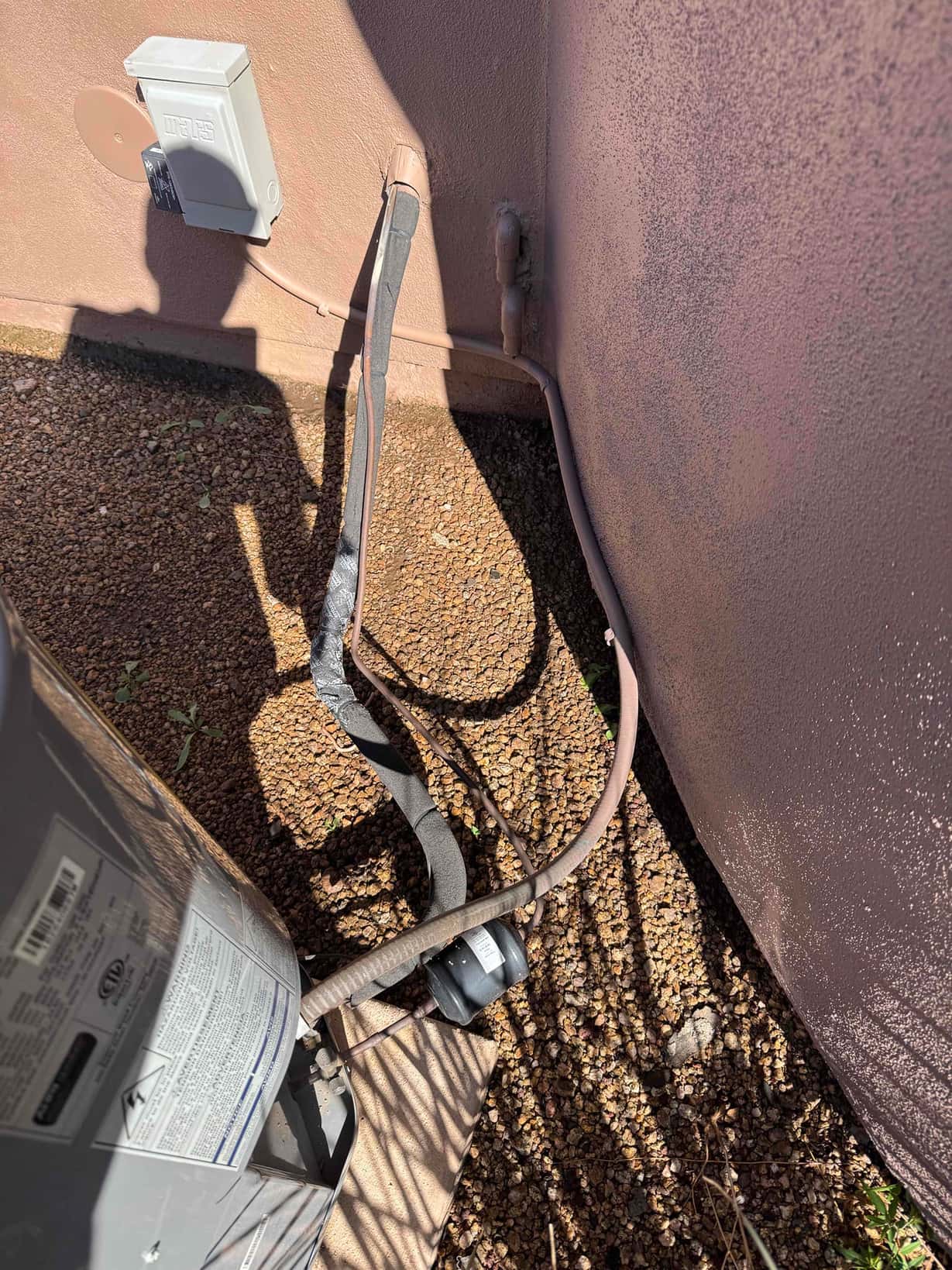 Outdoor condenser area showing refrigerant line set with deteriorated insulation, wall-mounted electrical disconnect, and line penetration at the wall.