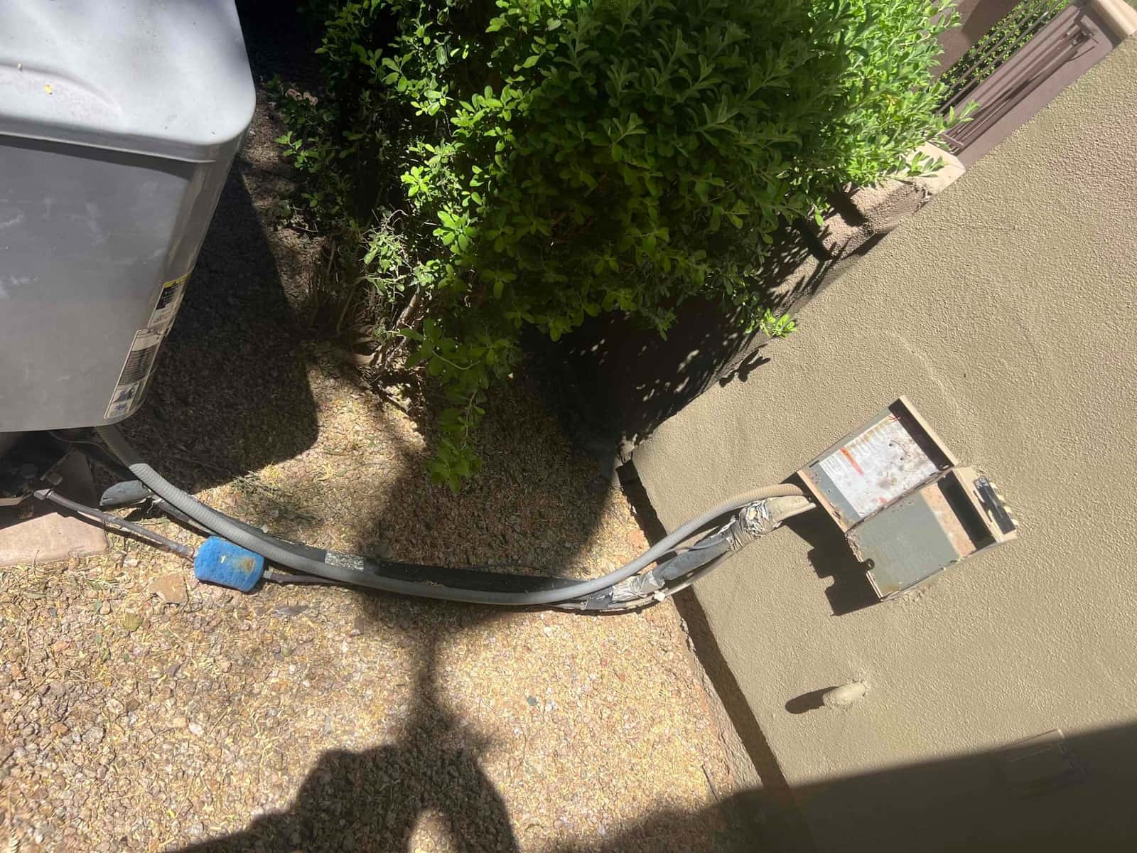 Outdoor condenser beside wall with open electrical disconnect, flexible conduit, and insulated refrigerant lines routed along gravel ground.