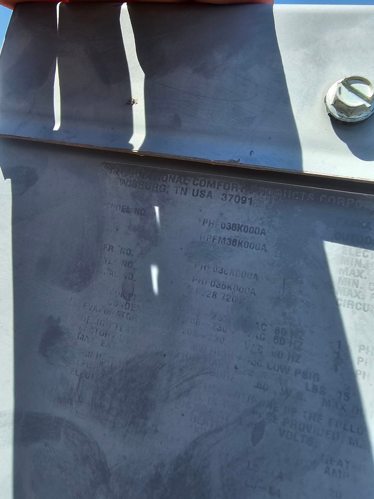 Outdoor condenser cabinet panel with a faded equipment data plate under sunlight and hand shadows.