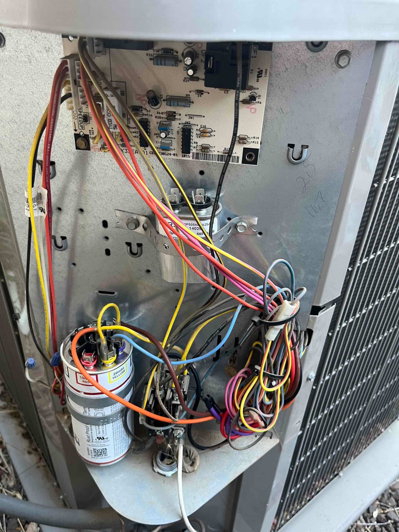 Outdoor condenser control panel open, showing dual-run capacitor, contactor, control board, and bundled low-voltage and line wiring.