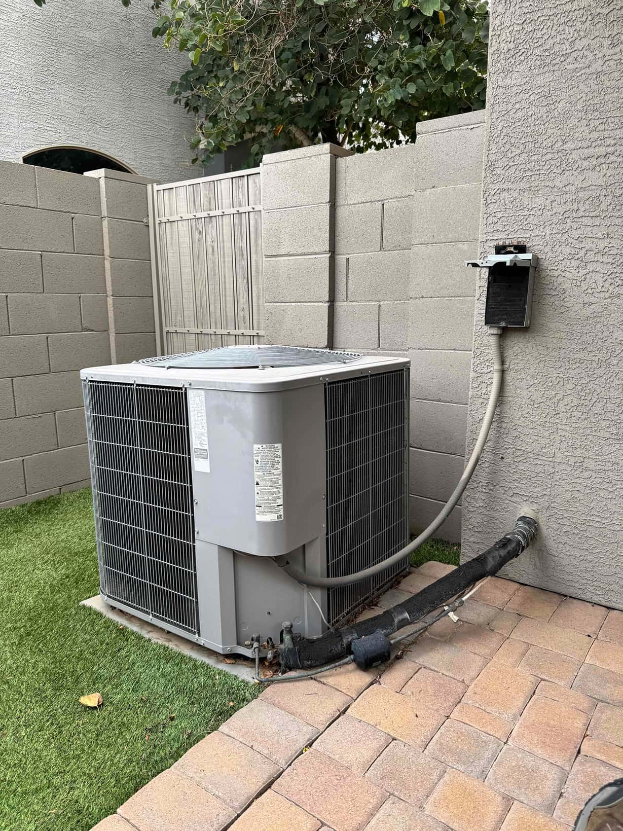 Outdoor condenser unit beside house with insulated refrigerant lines, service valves, and an open electrical disconnect mounted on the wall.