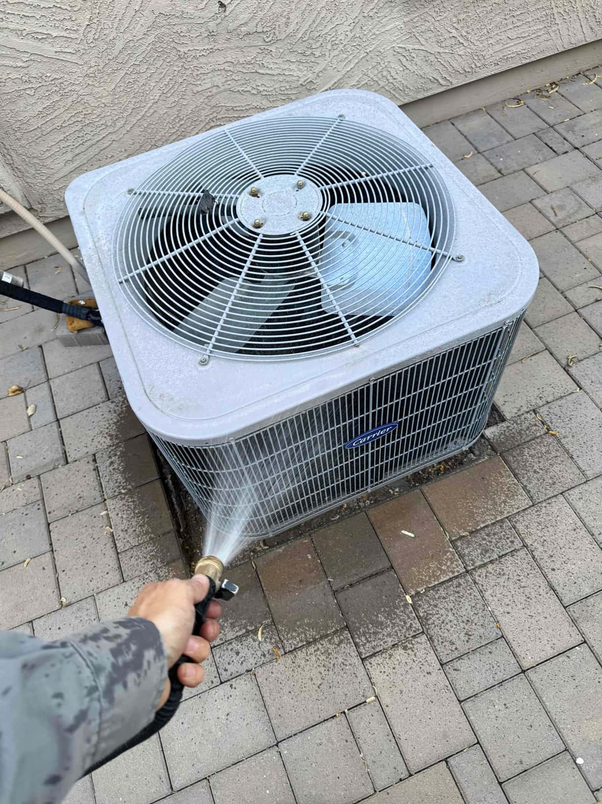 Outdoor condenser unit being rinsed with a hose, water spraying across coil fins and grille.