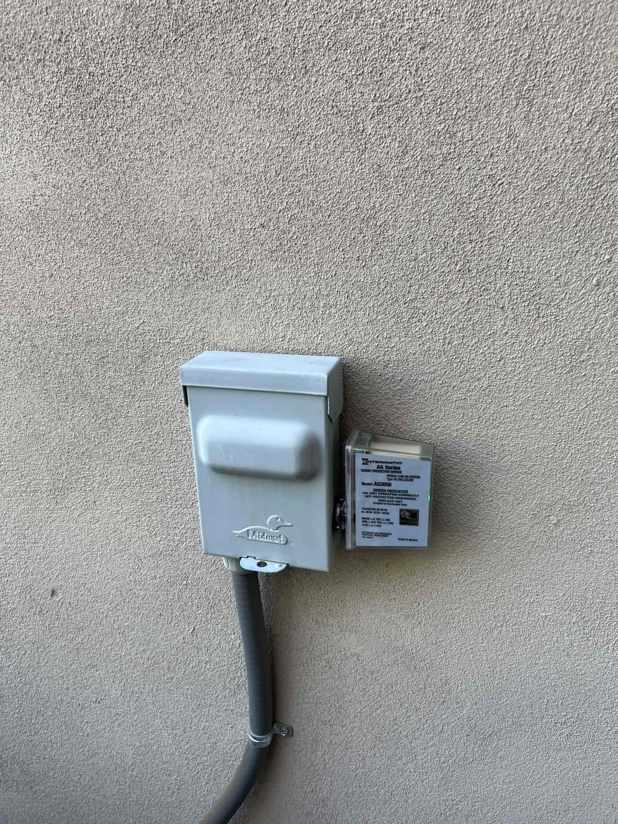 Outdoor HVAC service disconnect mounted on stucco wall with flexible conduit and adjacent protective device with illuminated status indicator.