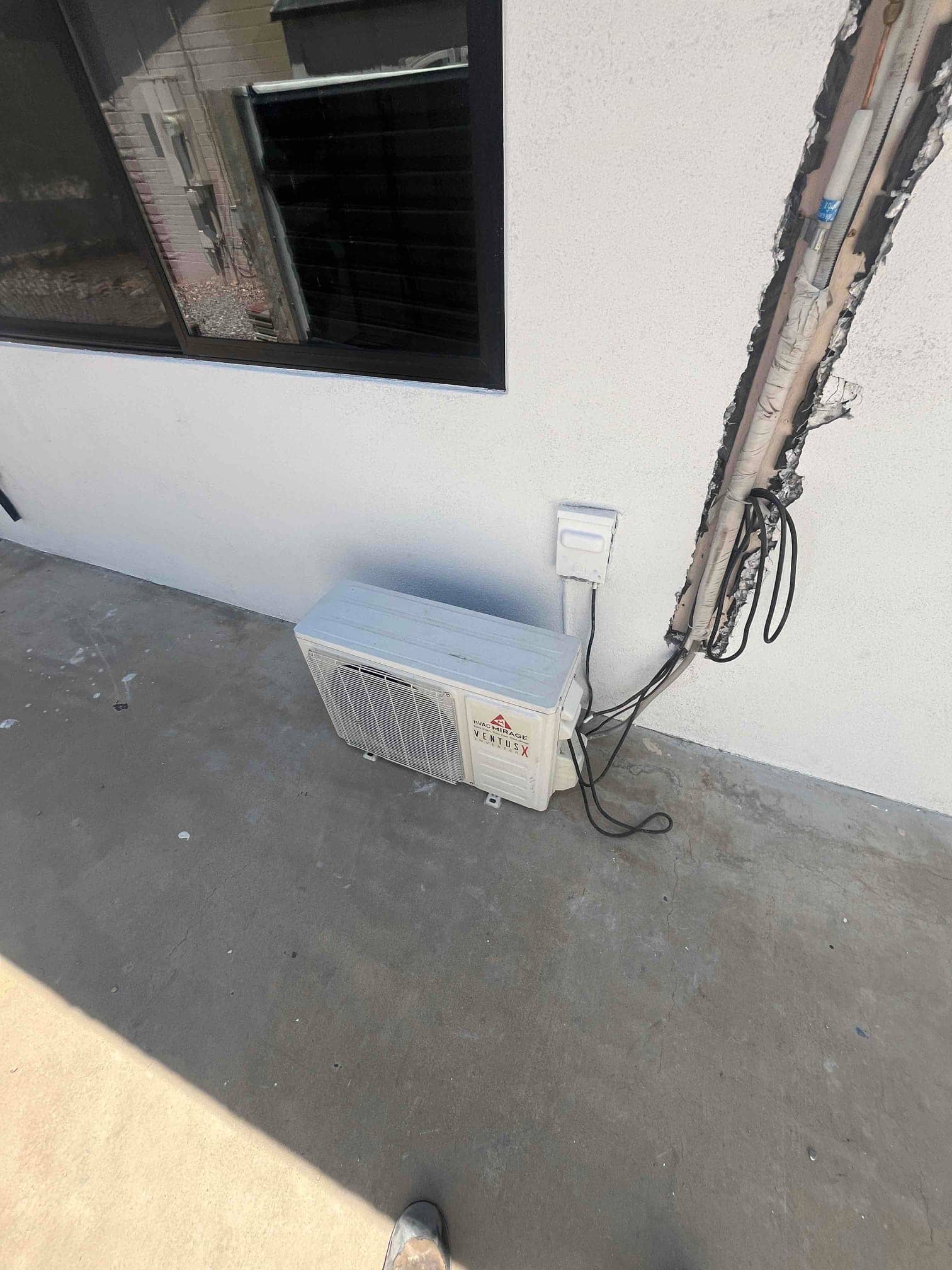 Outdoor mini-split condenser sits on concrete pad with open wall chase exposing insulated refrigerant lines and wiring.