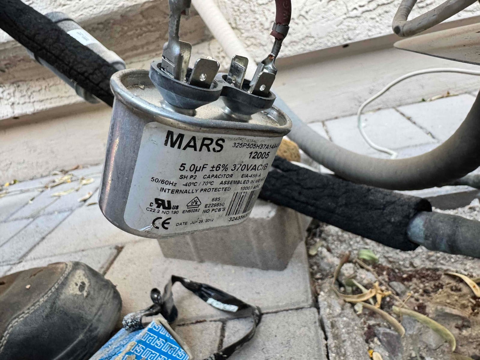 Oval run capacitor labeled 5.0 μF ±6% 370VAC/B with wiring connected to its terminals outdoors.