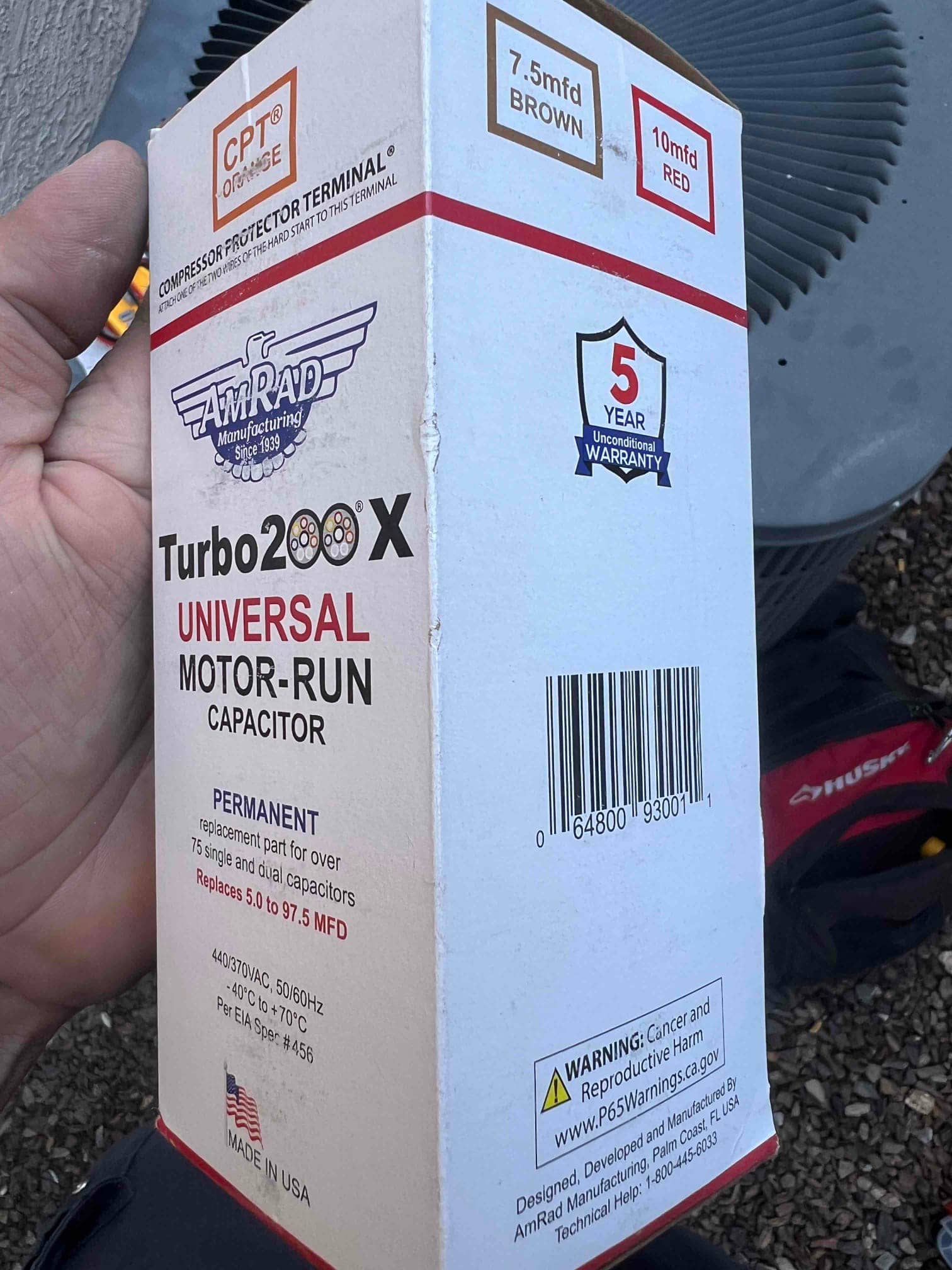 Packaging for a universal motor-run capacitor with printed specs, held beside an outdoor condenser during a residential service visit.