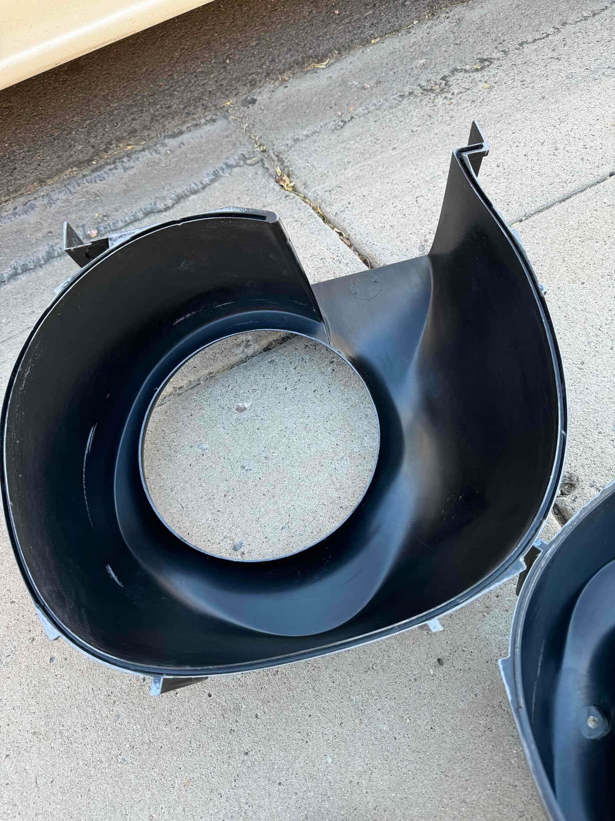Plastic fan shroud housing on the ground, circular center opening with visible mounting tabs and curved interior surfaces.
