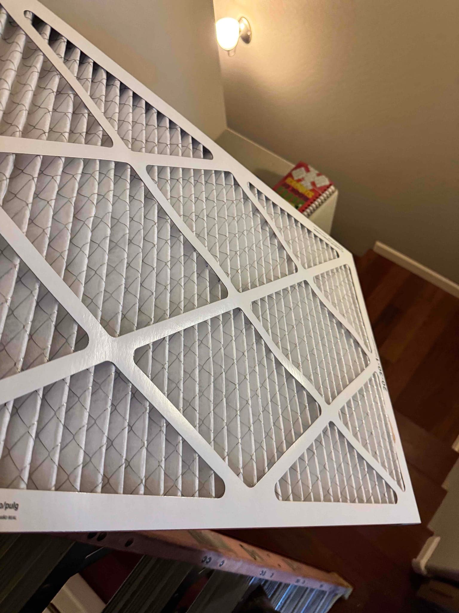 Pleated HVAC air filter with cardboard frame shown close-up, positioned indoors with a ladder underneath.