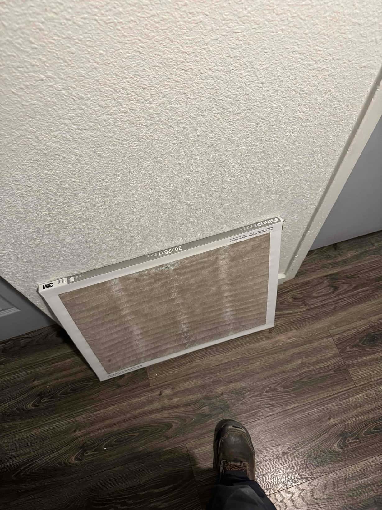 Pleated HVAC air filter leaning against a wall on laminate flooring near an interior doorway.