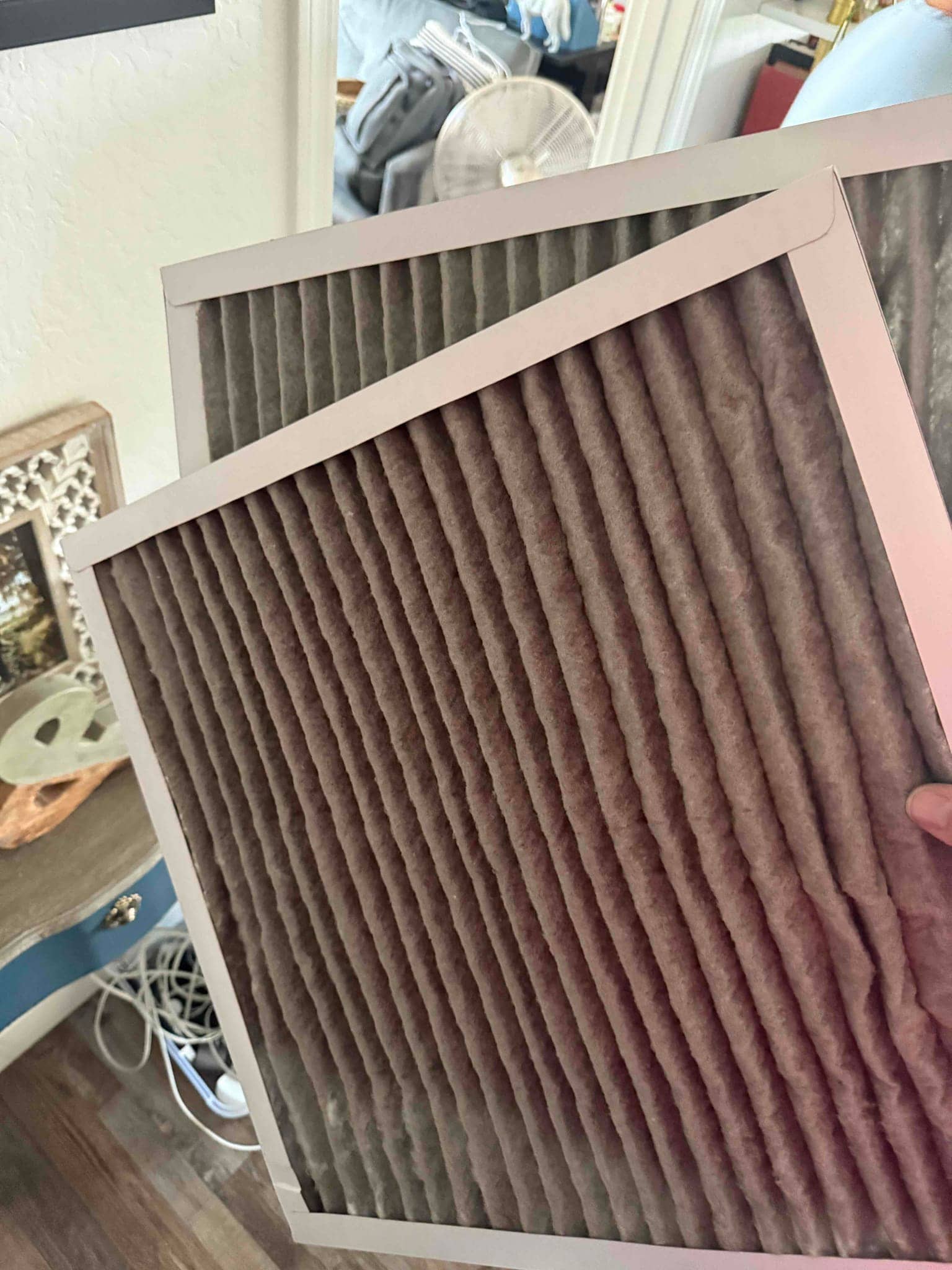 Two pleated HVAC air filters are held indoors, showing visible dust accumulation across the filter media.