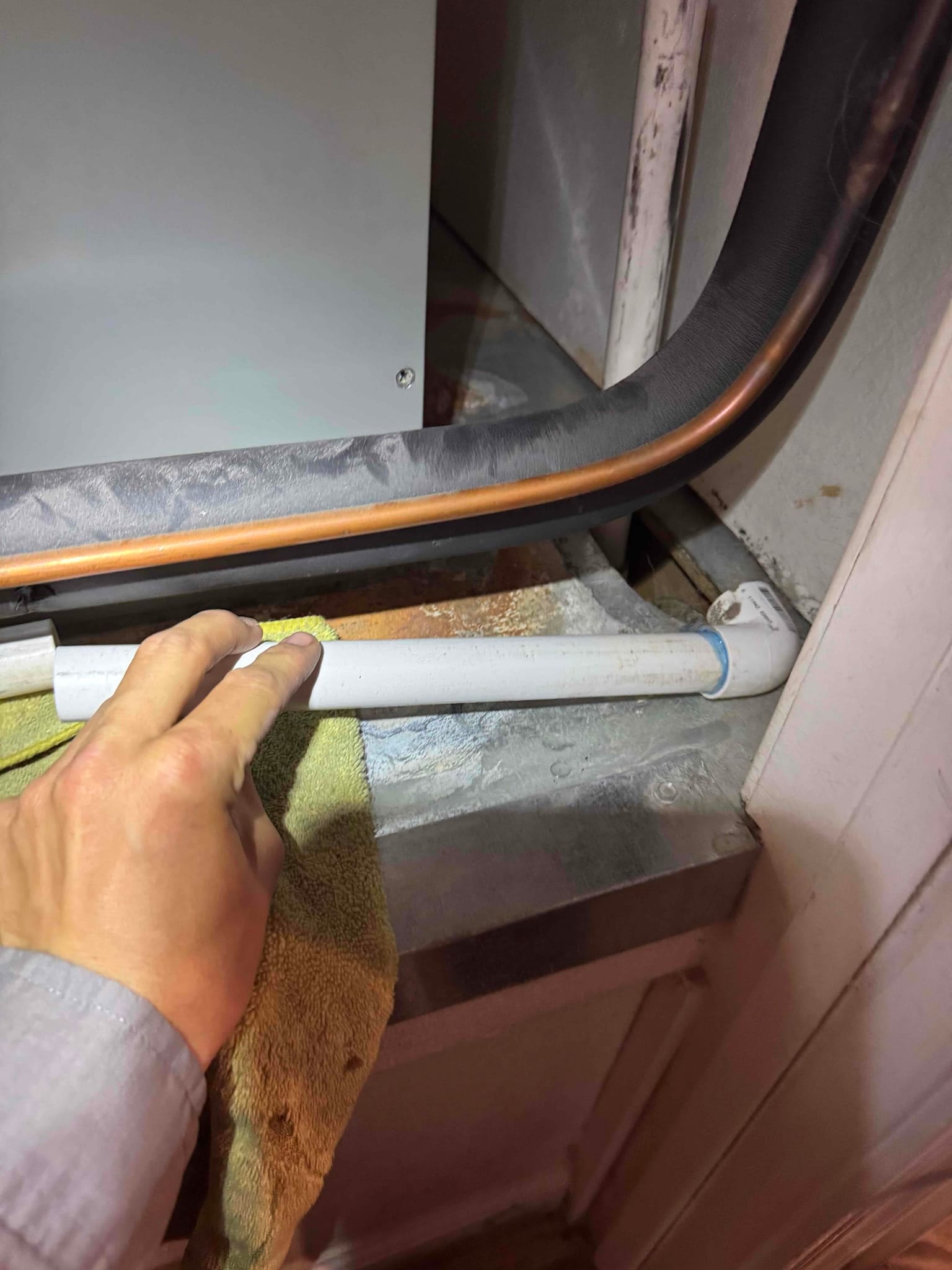 PVC condensate drain with elbow runs along metal pan beneath insulated copper refrigerant lines; visible rust staining at pan.