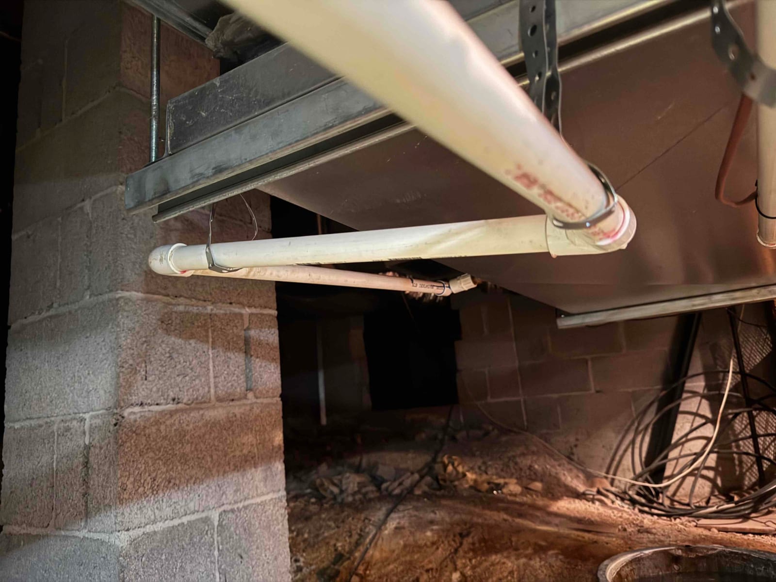 PVC condensate drain lines routed beneath an indoor unit in a crawlspace, secured with metal straps.